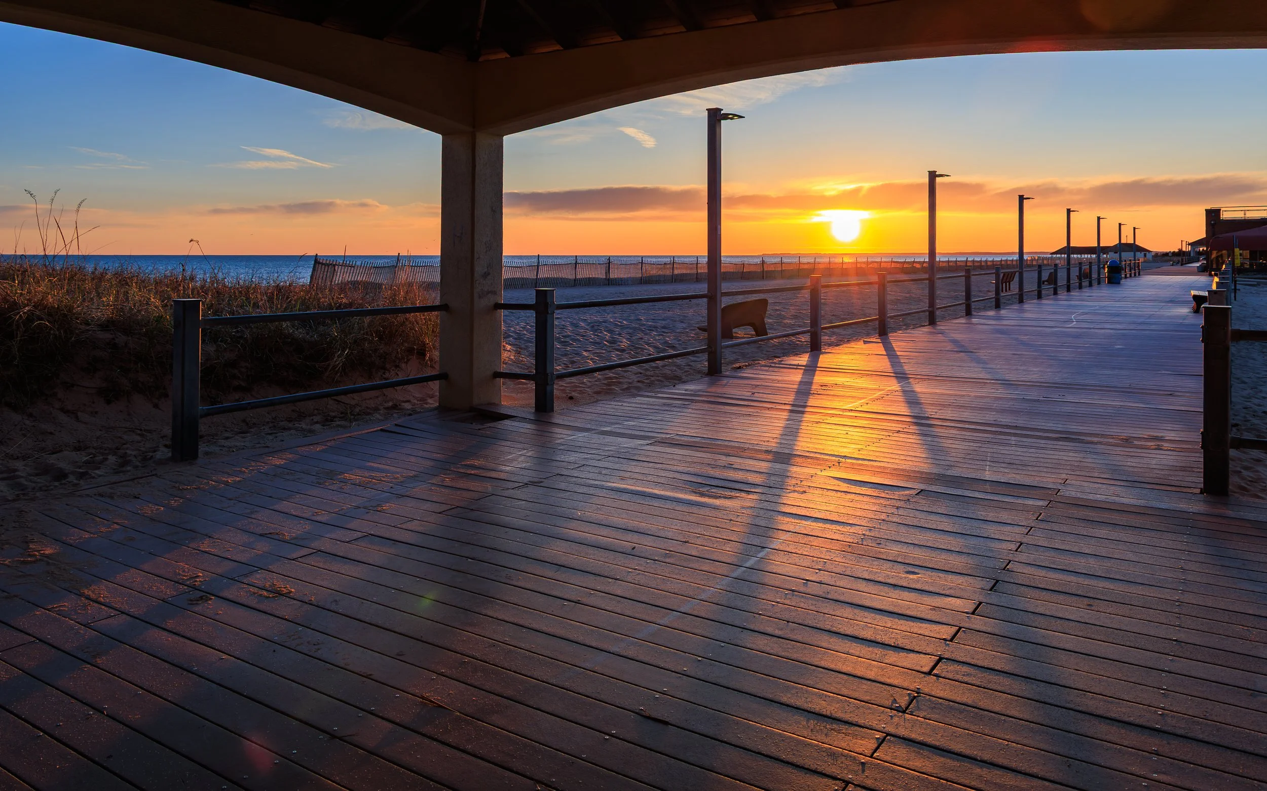 Sunset over a beach promenade with wooden deck flooring, benches, and street lamps, viewed from underneath a sheltered area.