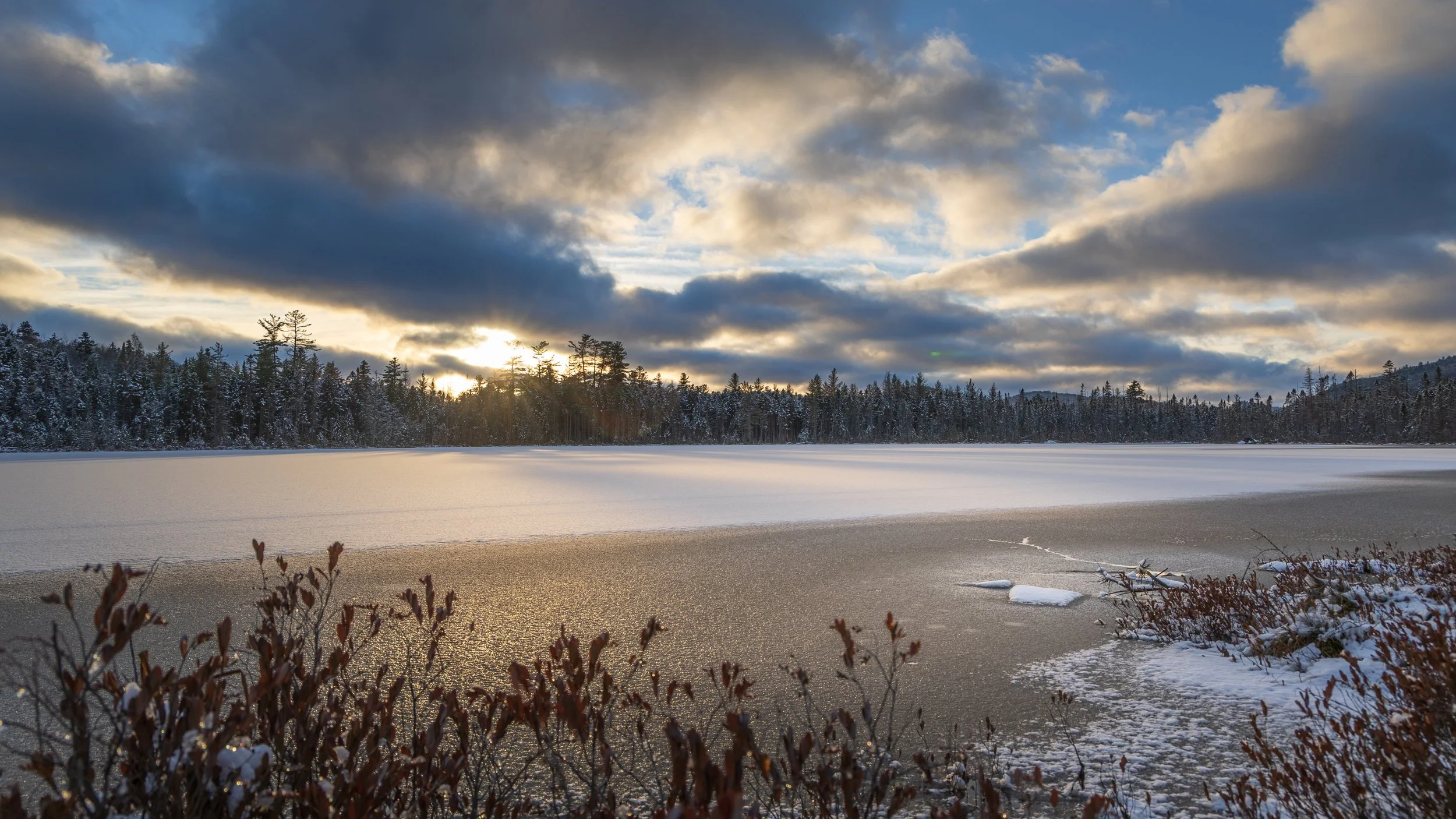 Frozen lake with snow-covered edges, surrounded by a forest of snow-covered trees, under a cloudy sky with sunlight breaking through.