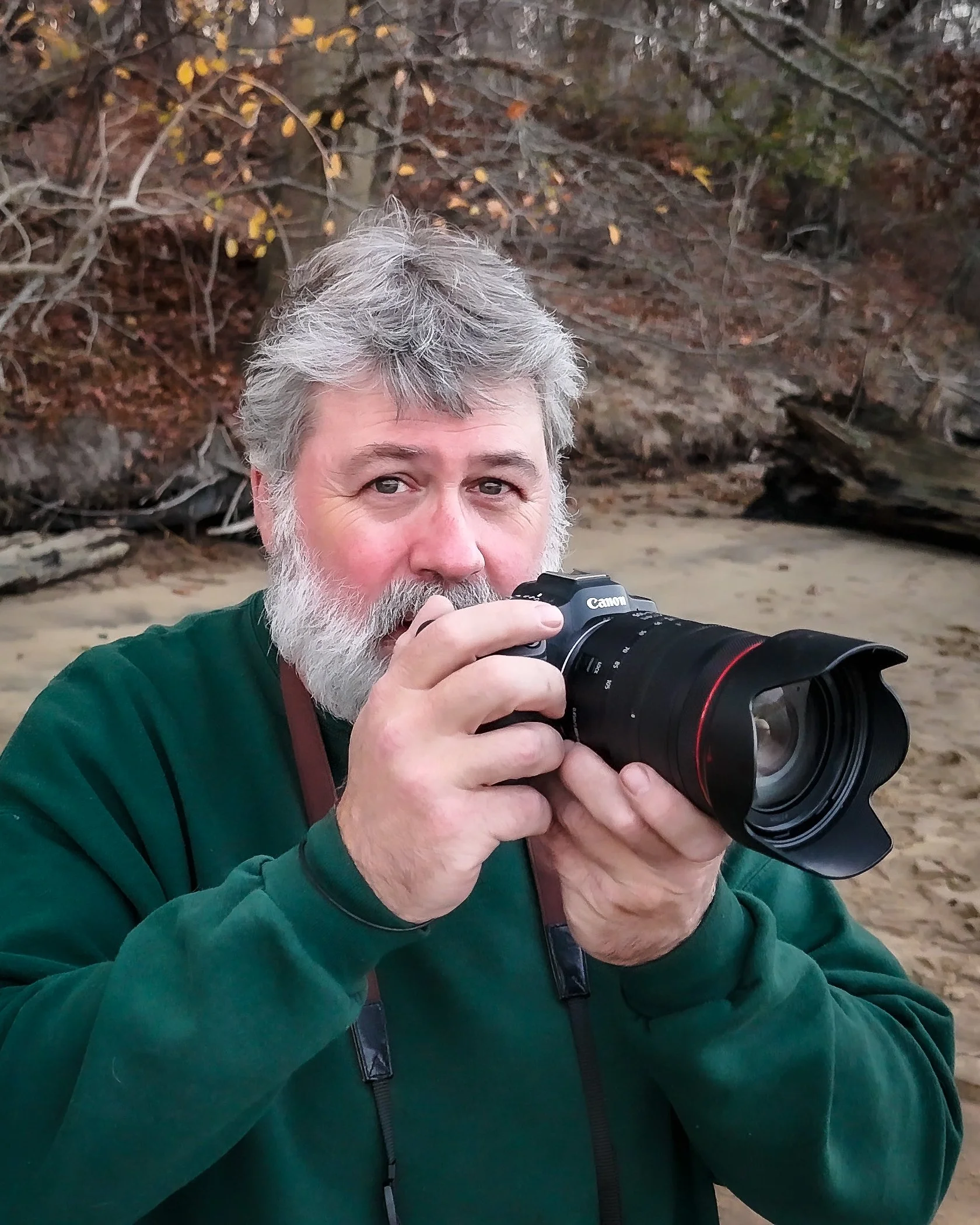 A man with gray hair and a beard is holding a Canon camera, looking through the viewfinder at an outdoor setting with trees in the background.