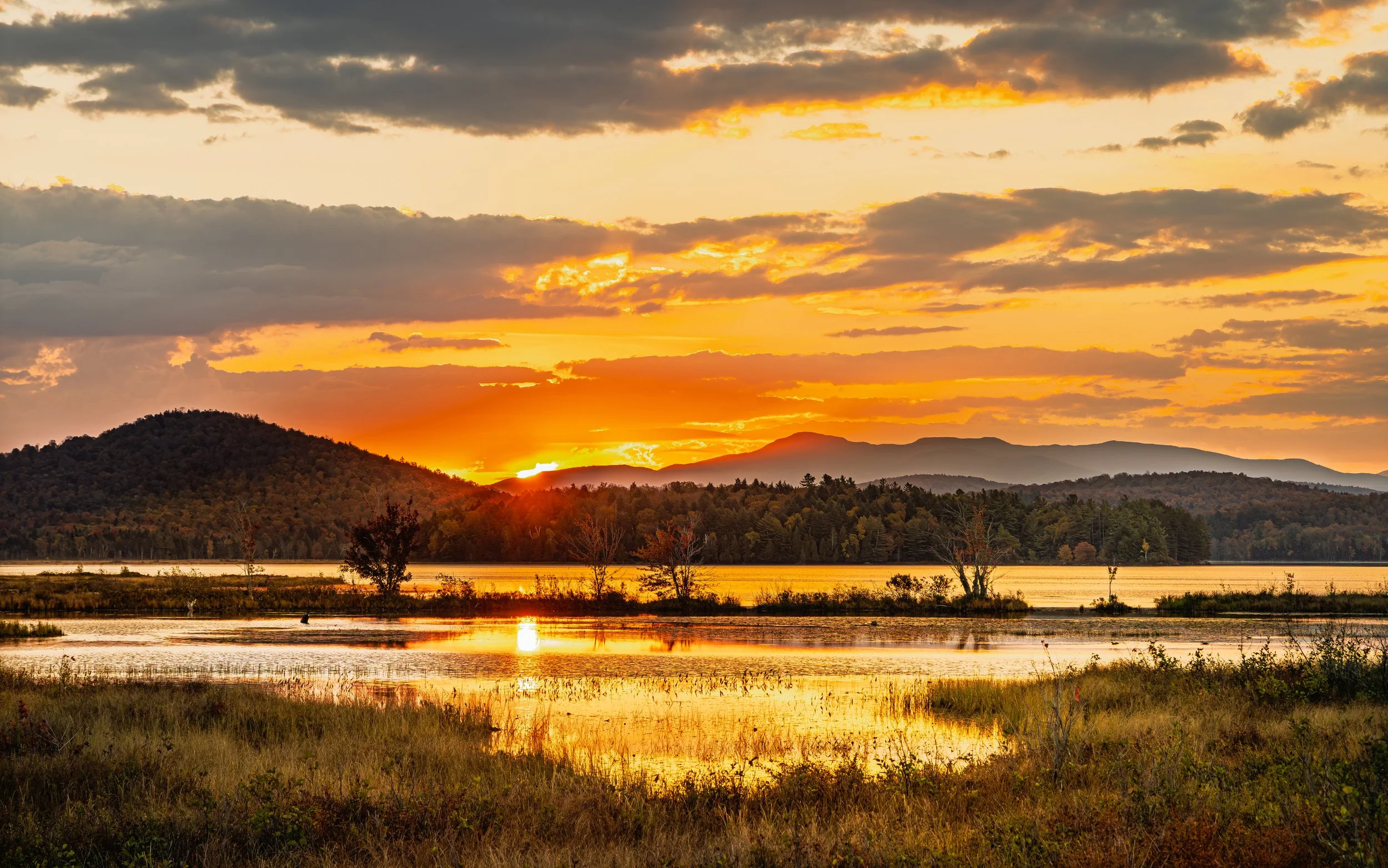 A scenic view of a sunset over mountains and a body of water with trees and grassy area in the foreground.