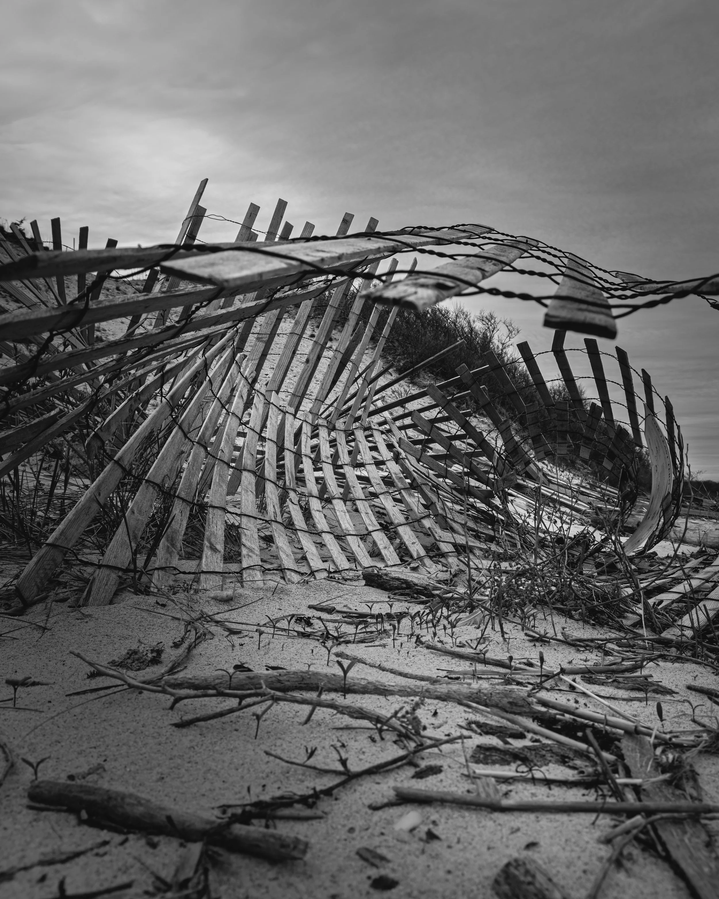 A black and white photograph of a damaged wooden and wire fence on a sandy terrain. The fence appears bent and broken, with some parts leaning or fallen over. The sky above is cloudy, and there are some trees or shrubs in the background.