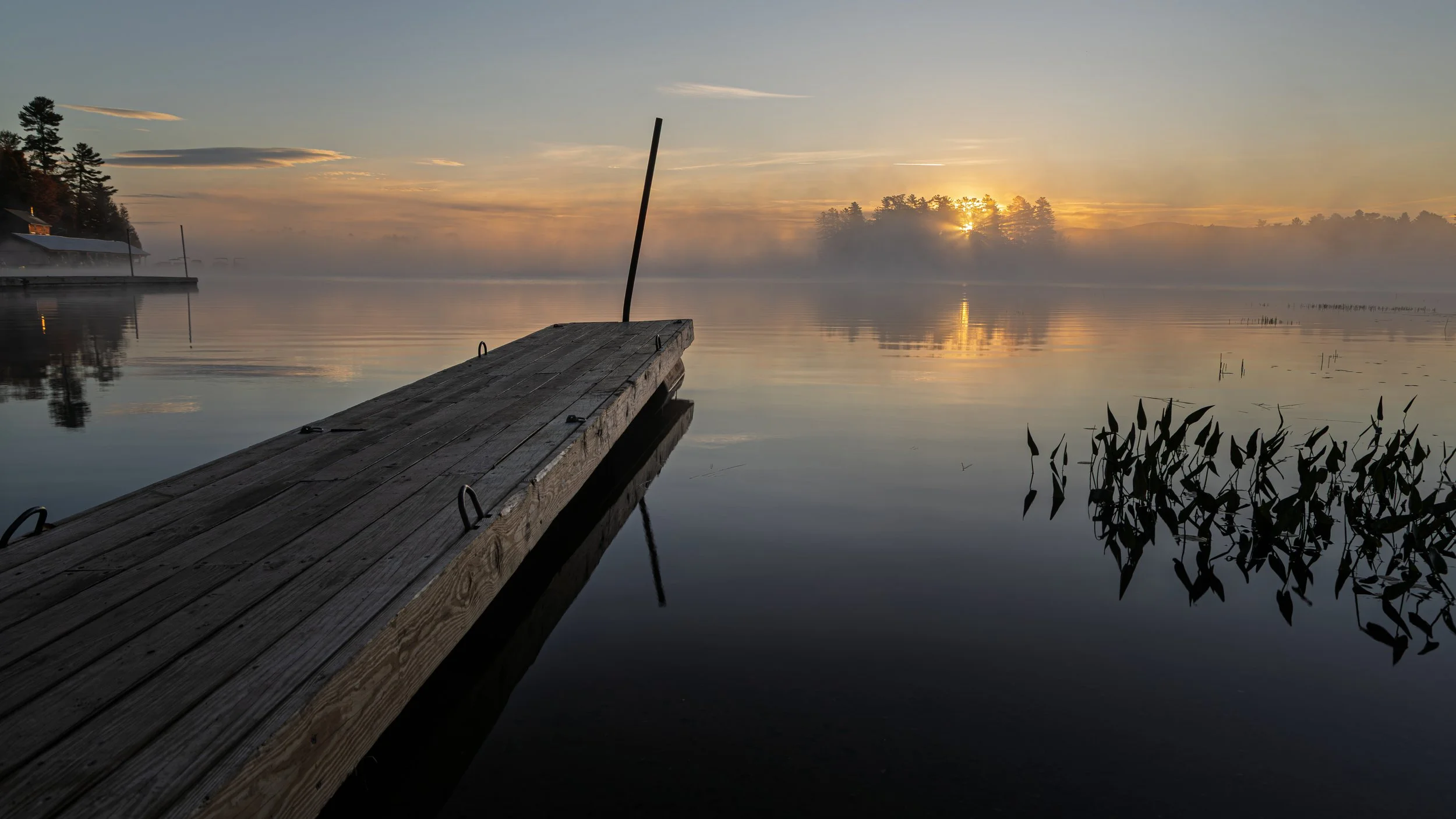 A wooden dock extending into a calm lake at sunrise, with mist hovering above the water and trees silhouetted in the background.