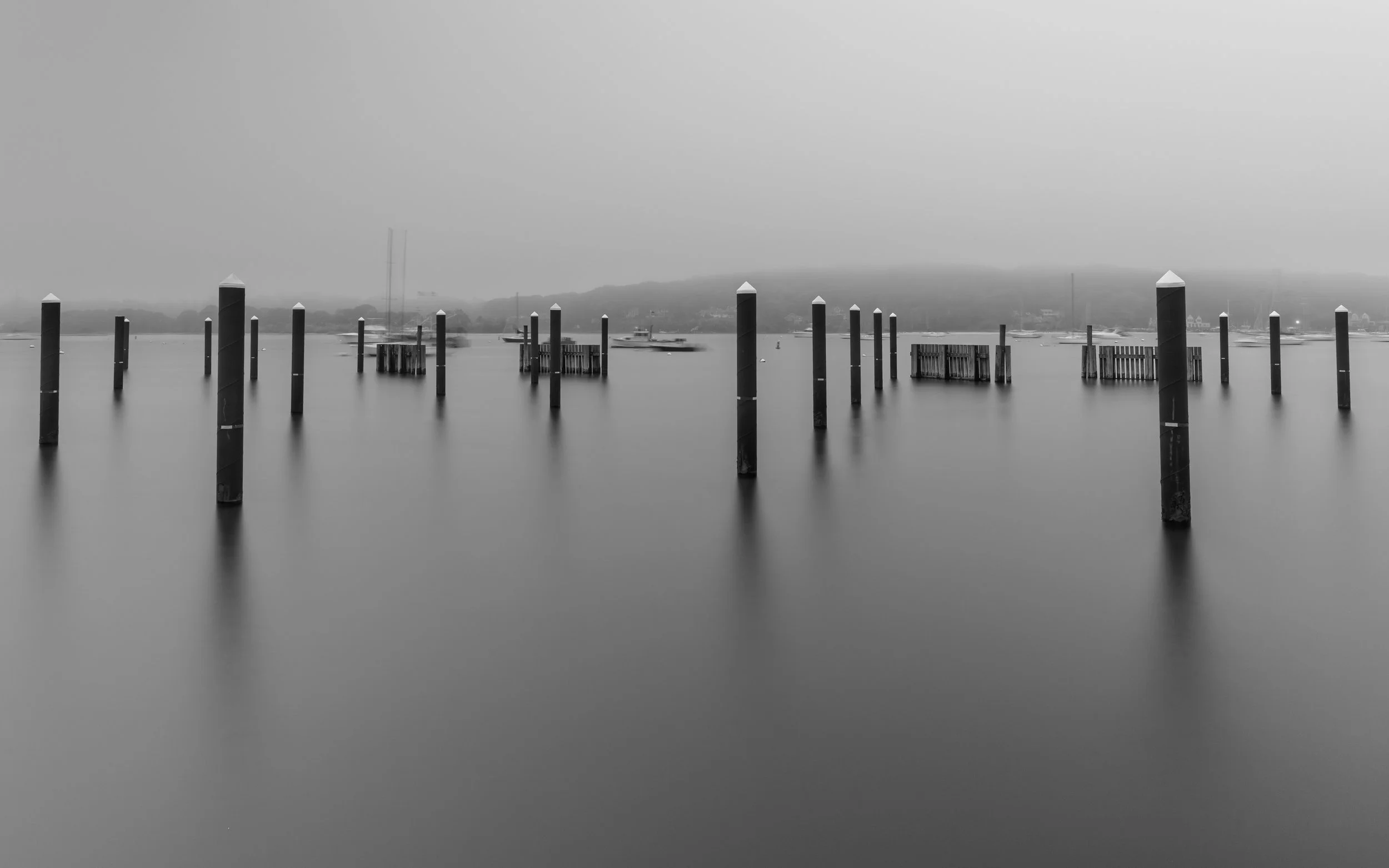 Black and white photograph of a calm body of water with several wooden pilings and small docks, with a misty shoreline in the background.