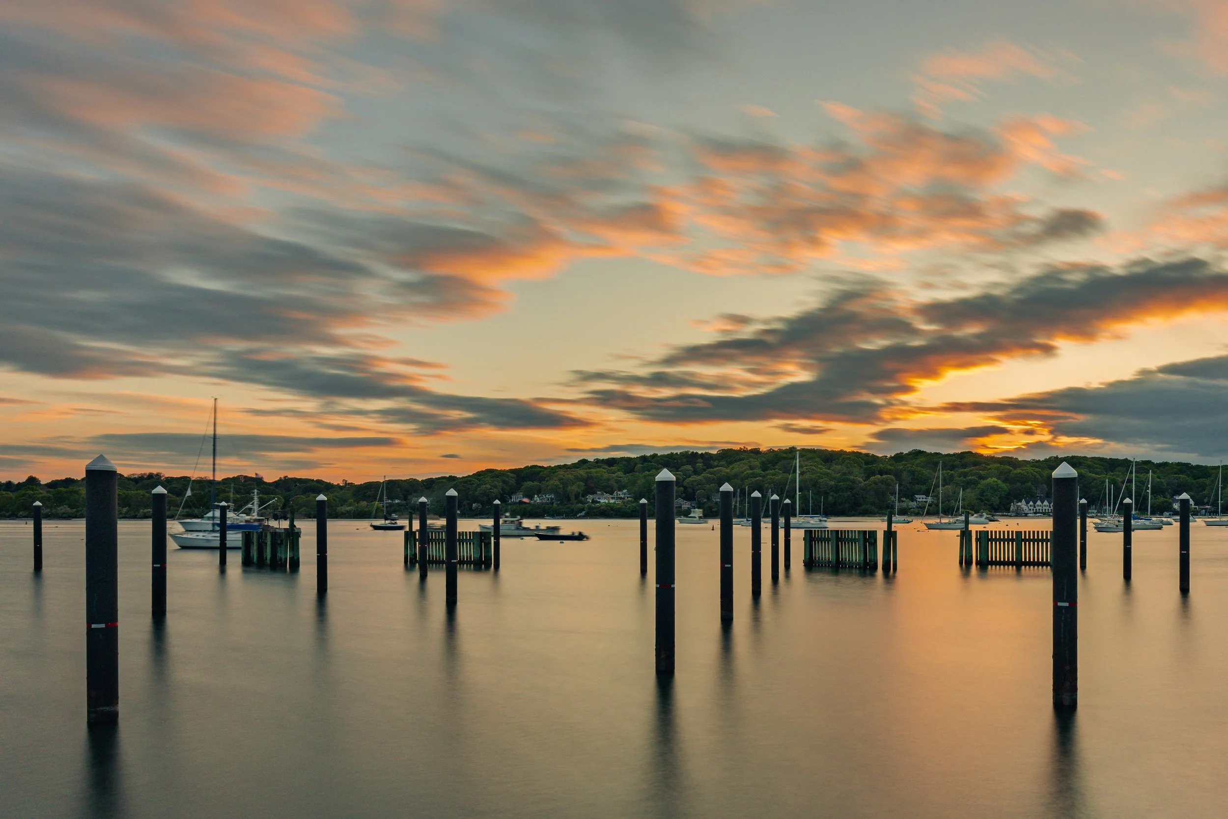 A marina at sunset with sailboats and motorboats anchored, wooden docks, and pilings in calm water. The sky has colorful clouds with shades of orange, pink, and blue, with hills and trees in the background.