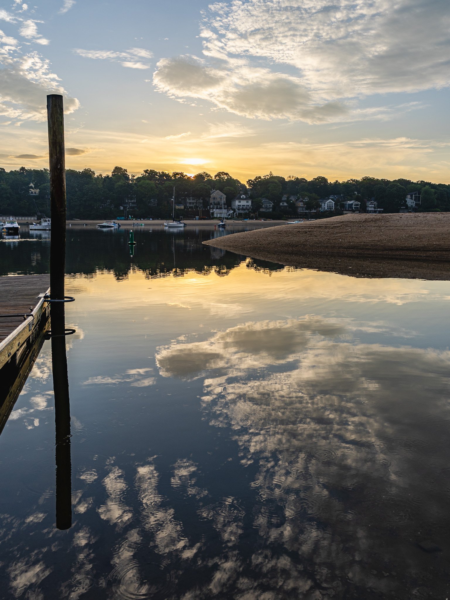 Sunset over a calm body of water with boats, houses on a hillside, and partly cloudy sky reflected in the water.