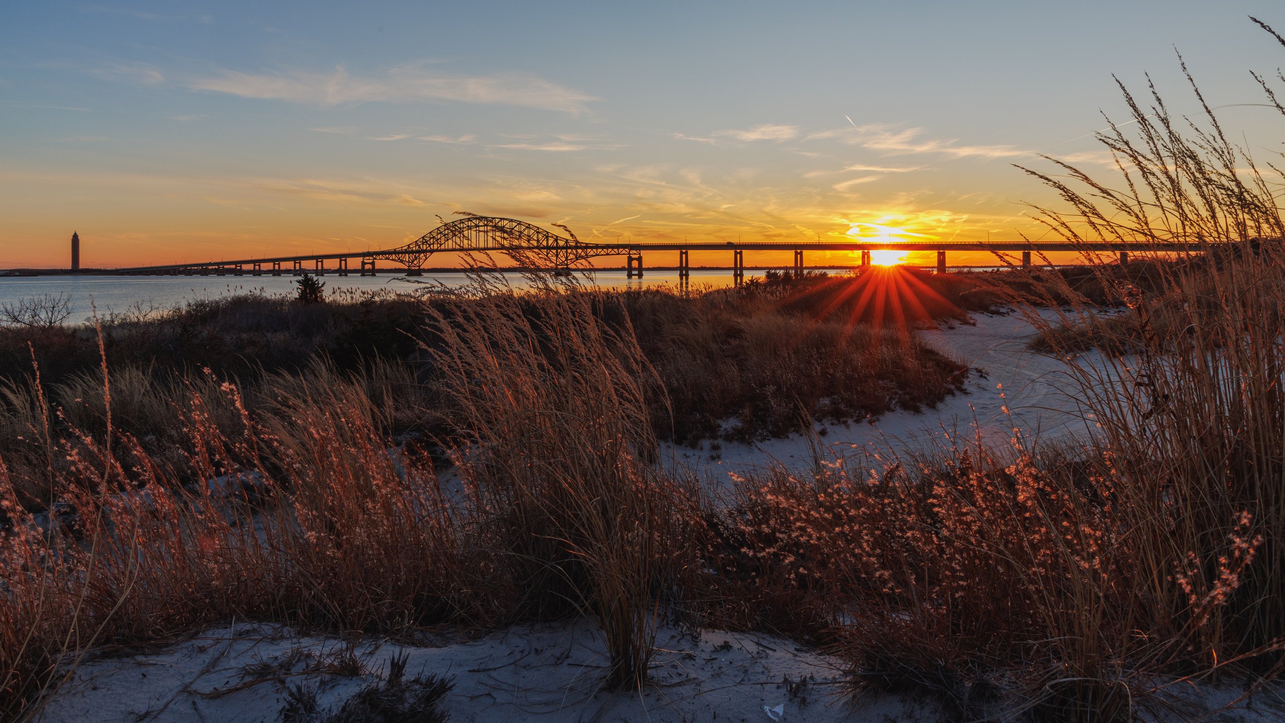 Sunset over a bridge spanning a body of water, with tall grasses and sand in the foreground.