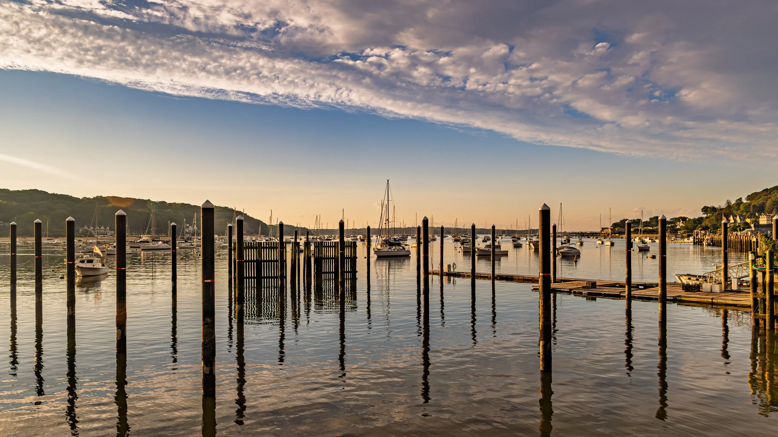 A serene harbor with calm water reflecting boats and wooden posts, with a hillside and houses in the background under a partly cloudy sky during sunset.