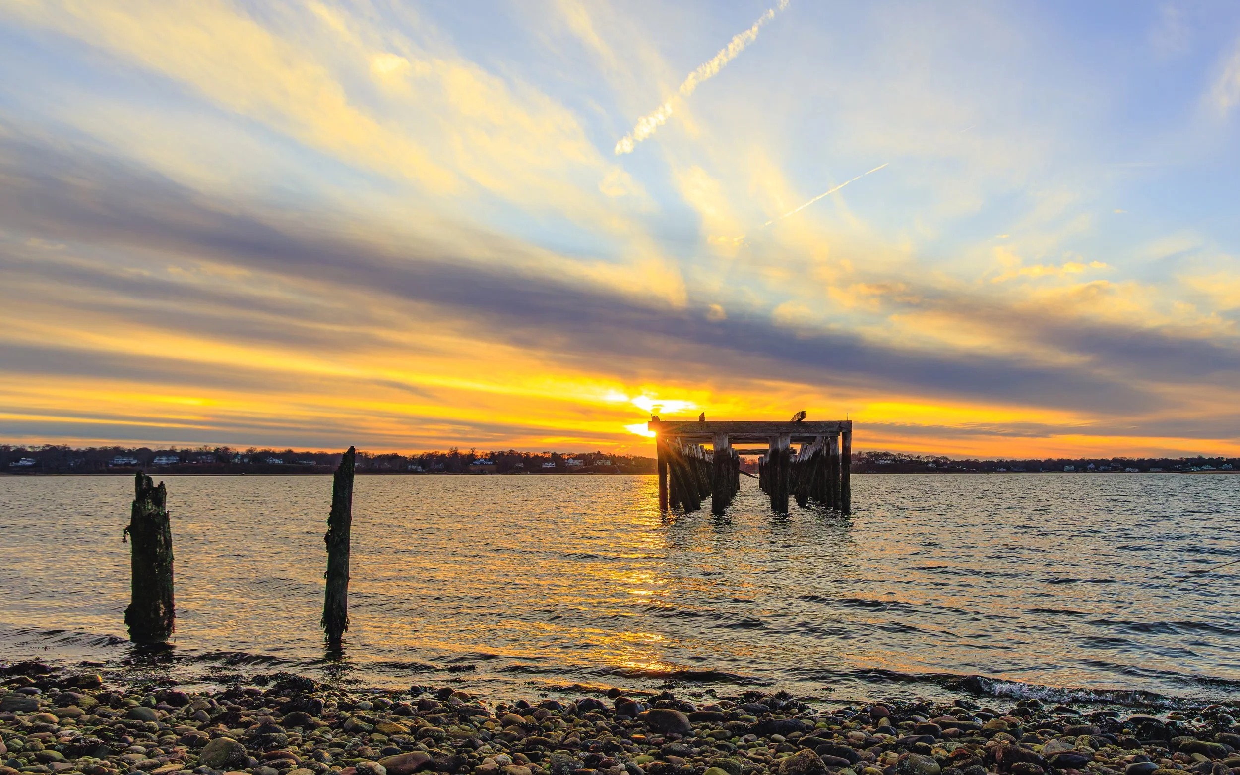 A sunset over a body of water with a cloudy sky and an old wooden pier extending into the water. The shoreline is covered with rocks, and the sky has streaks of clouds and contrails.