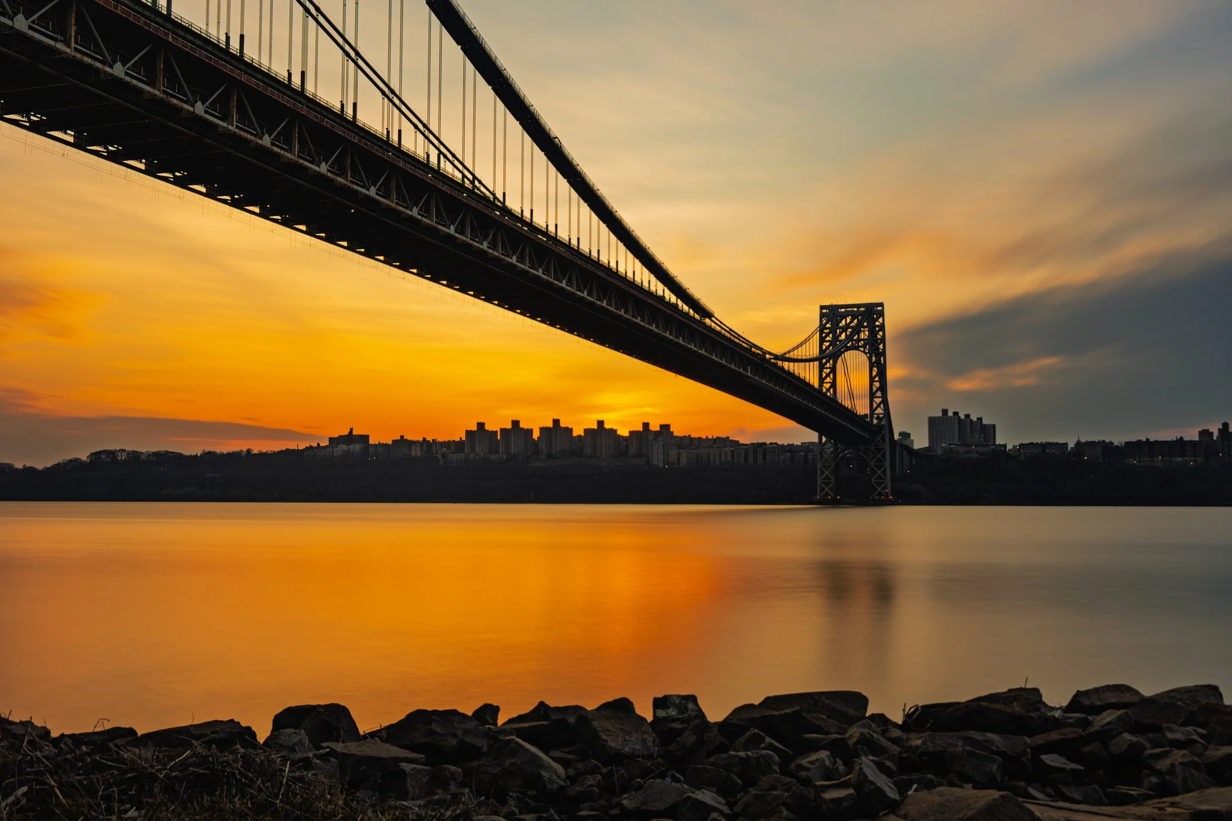 A large bridge extending over a body of water during sunset, with a city skyline in the background and rocks along the shoreline in the foreground.