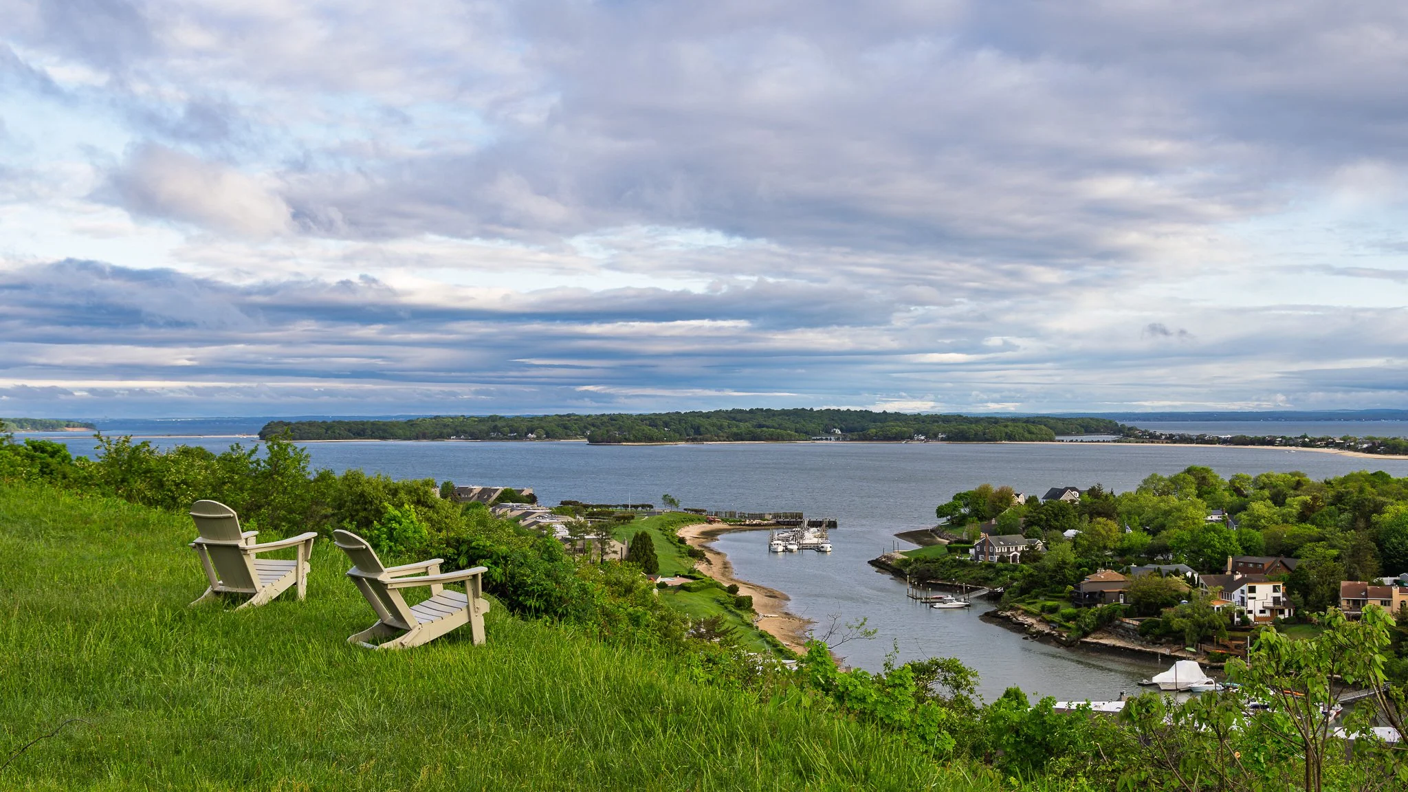 A scenic view of a river with houses along the shoreline, green trees, and a cloudy sky.