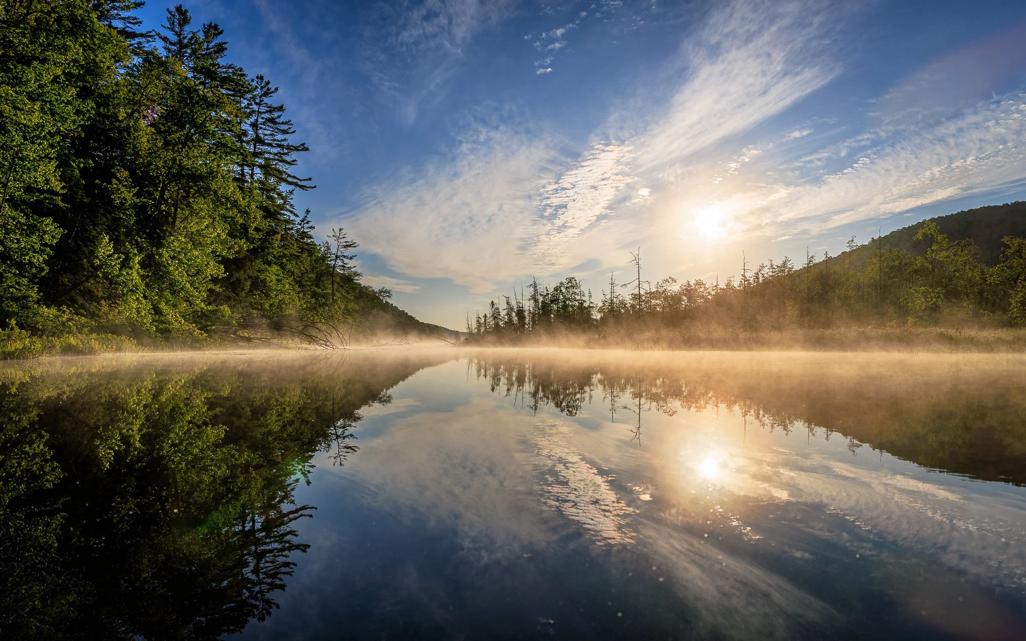 Morning fog over a calm river reflecting the grassy and wooded hills on either side, with a partly cloudy sky and the sun rising.