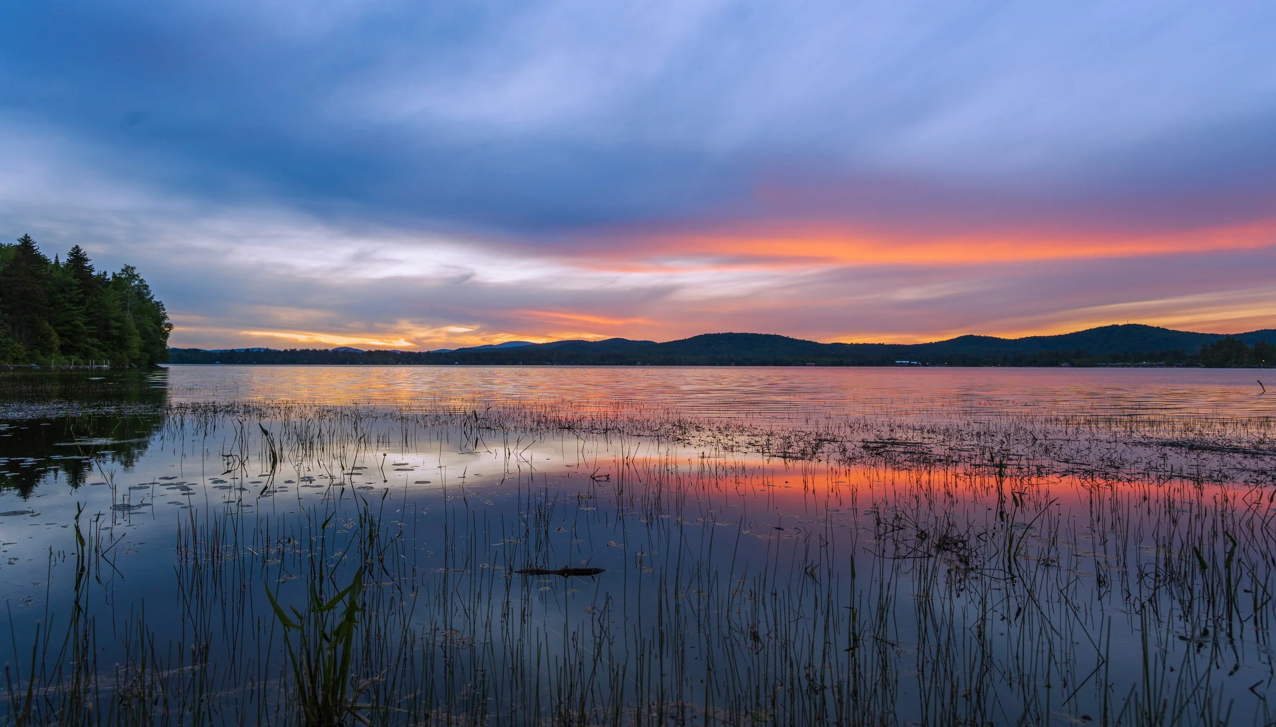 Sunset over a calm lake with reflections of colorful sky, trees on the shore, and distant hills.
