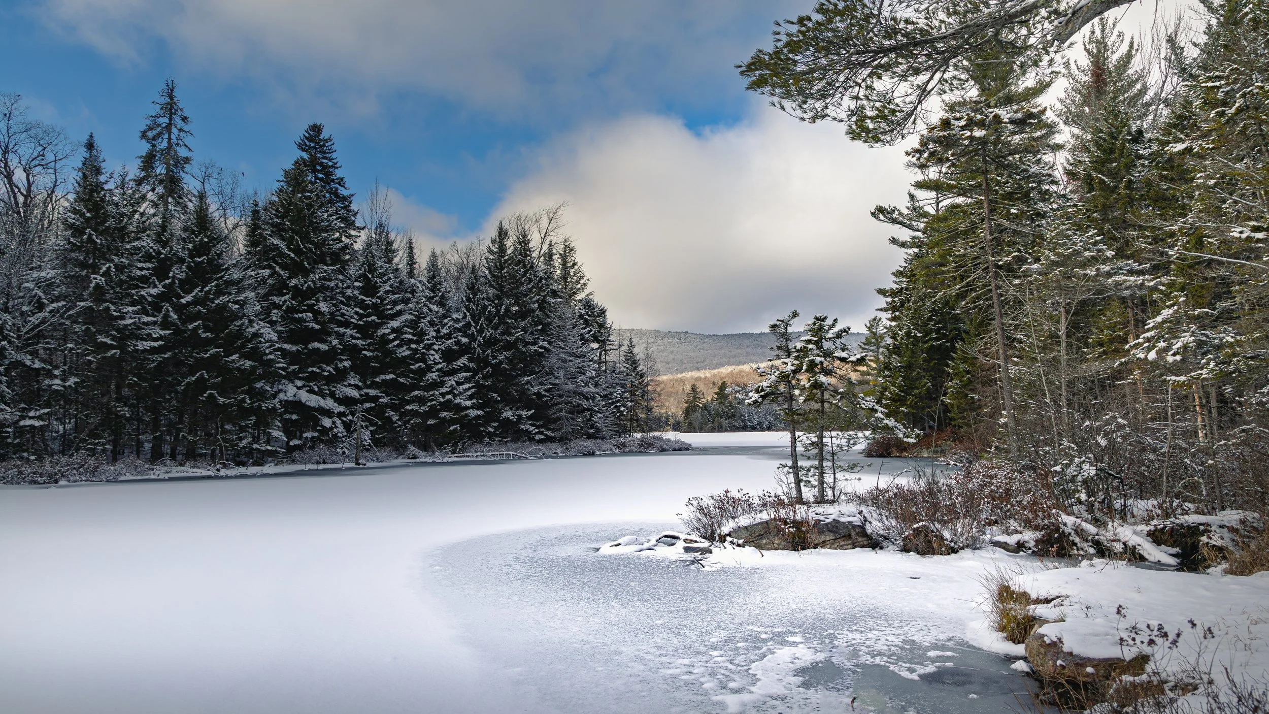 Snow-covered frozen lake surrounded by evergreen trees with snow on their branches, and a partially cloudy sky.