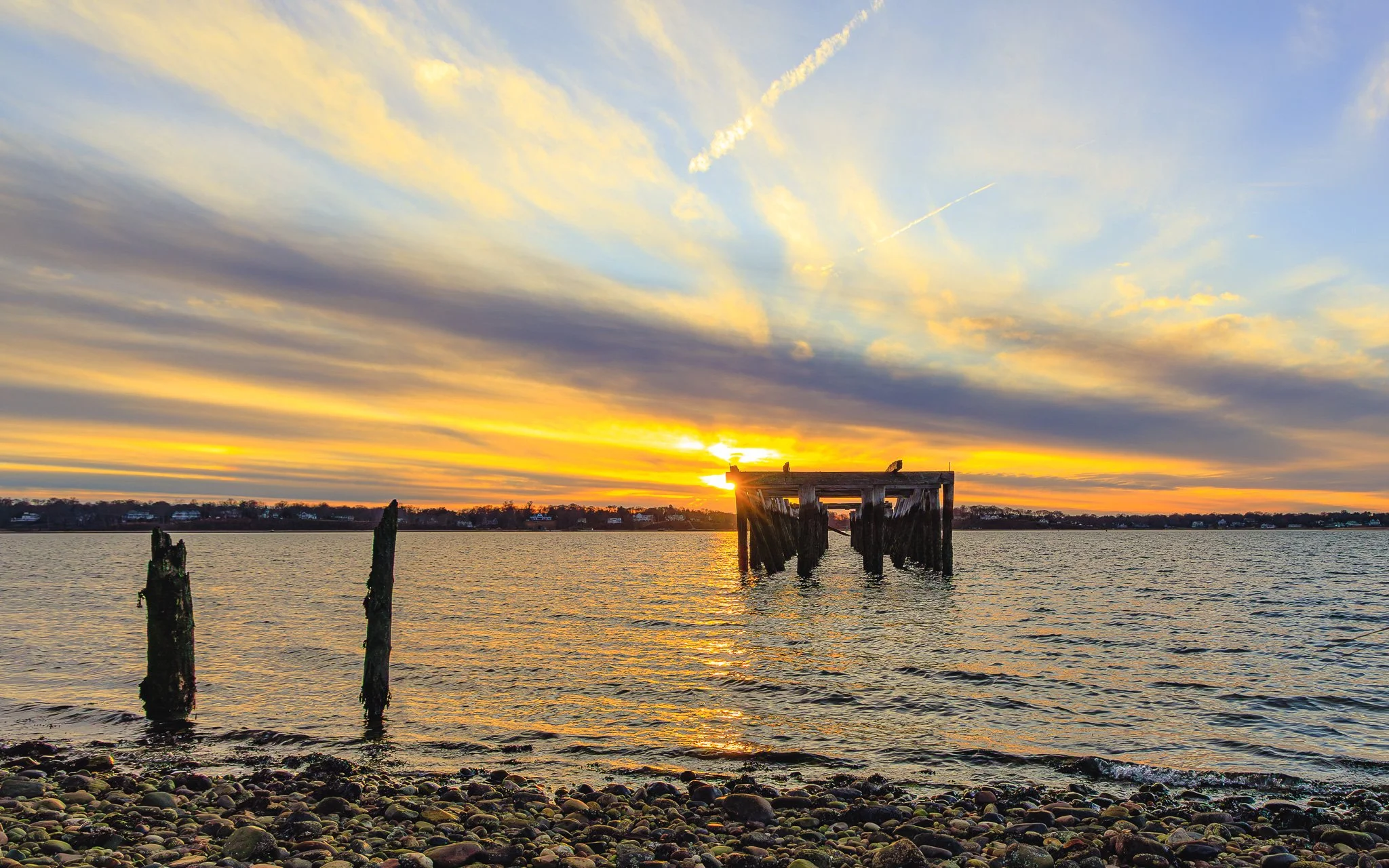 Sunset over a body of water with remnants of an old pier in the foreground and a distant tree line. The sky has streaks of yellow and blue clouds.