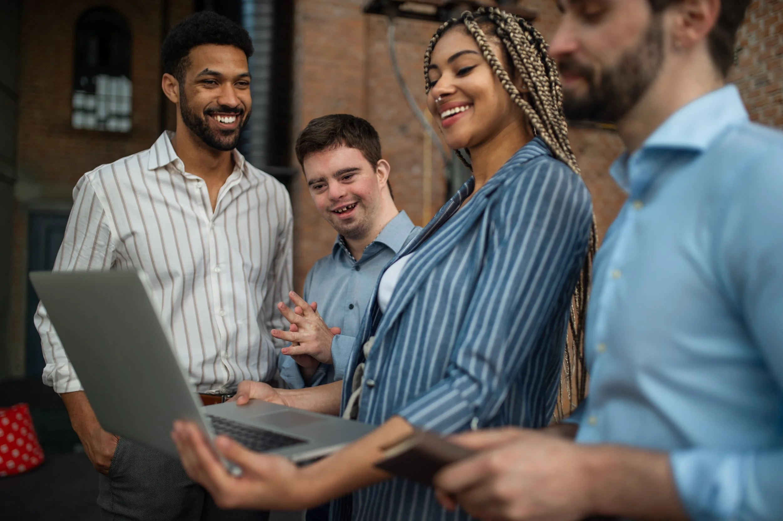 Three colleagues smile and gather around a laptop in a workplace setting. One team member, who has Down syndrome, is engaged and smiling as they look at the screen together.