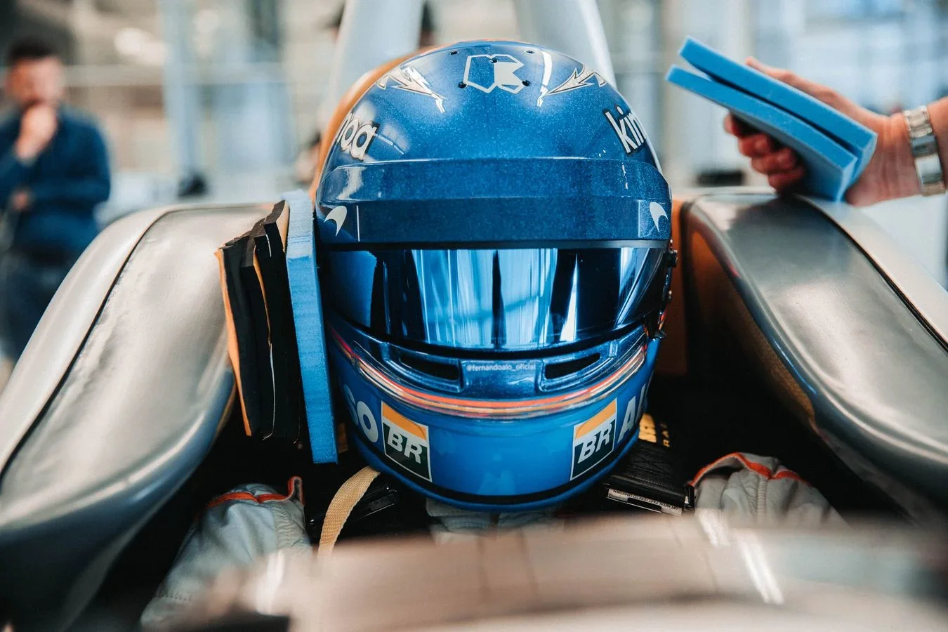 Close-up view of a blue racing helmet placed on a race car with garage staff in the background.