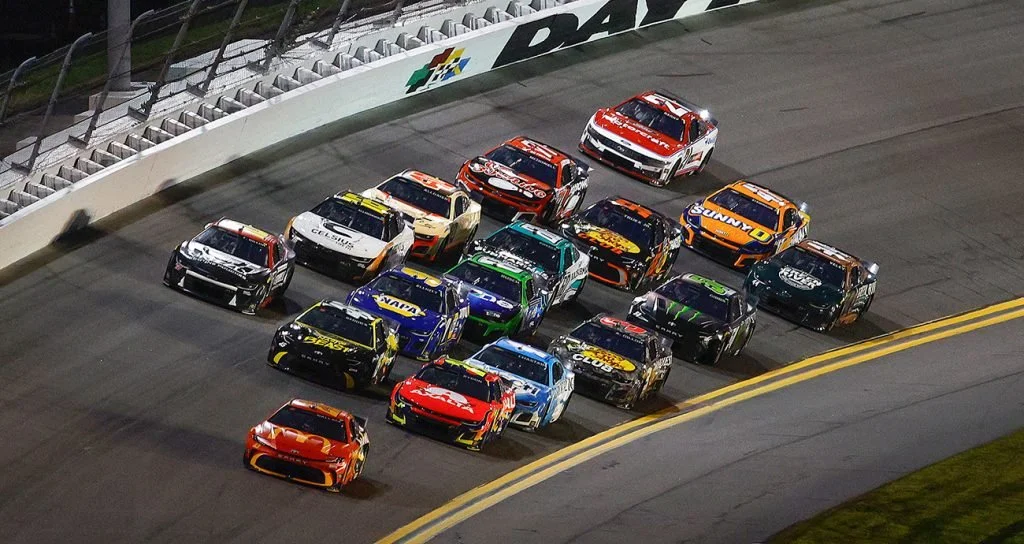 Multiple race cars on a track, likely during a NASCAR race, approaching a turn with a white barrier wall on the left and a yellow-lined curve on the right.