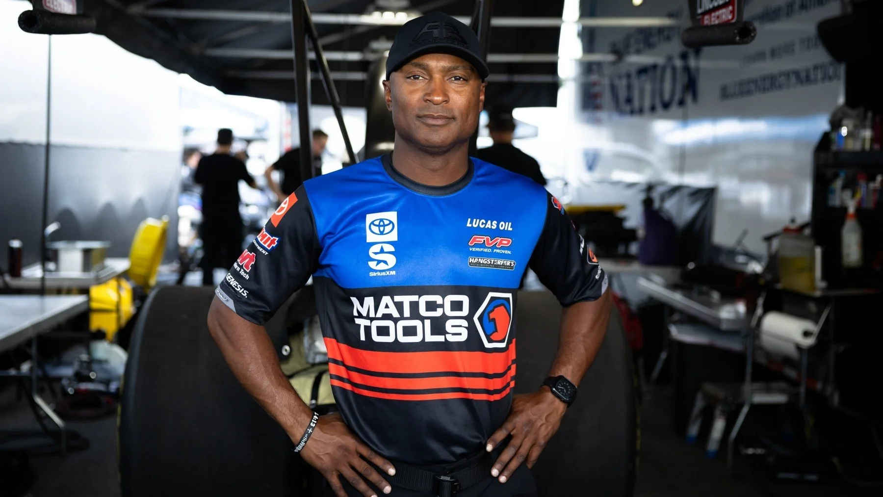 NHRA Top fuel driver Antron wearing a blue and black racing team uniform with various sponsor logos, standing in a garage area with equipment and team members in the background.