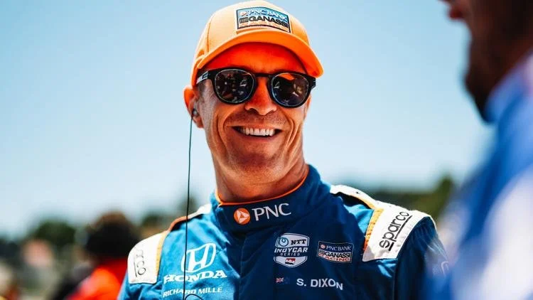 Indycar driver Scott Dixon Smiling man in racing helmet and sunglasses, wearing a blue racing suit with sponsor logos, outdoors against a blue sky.
