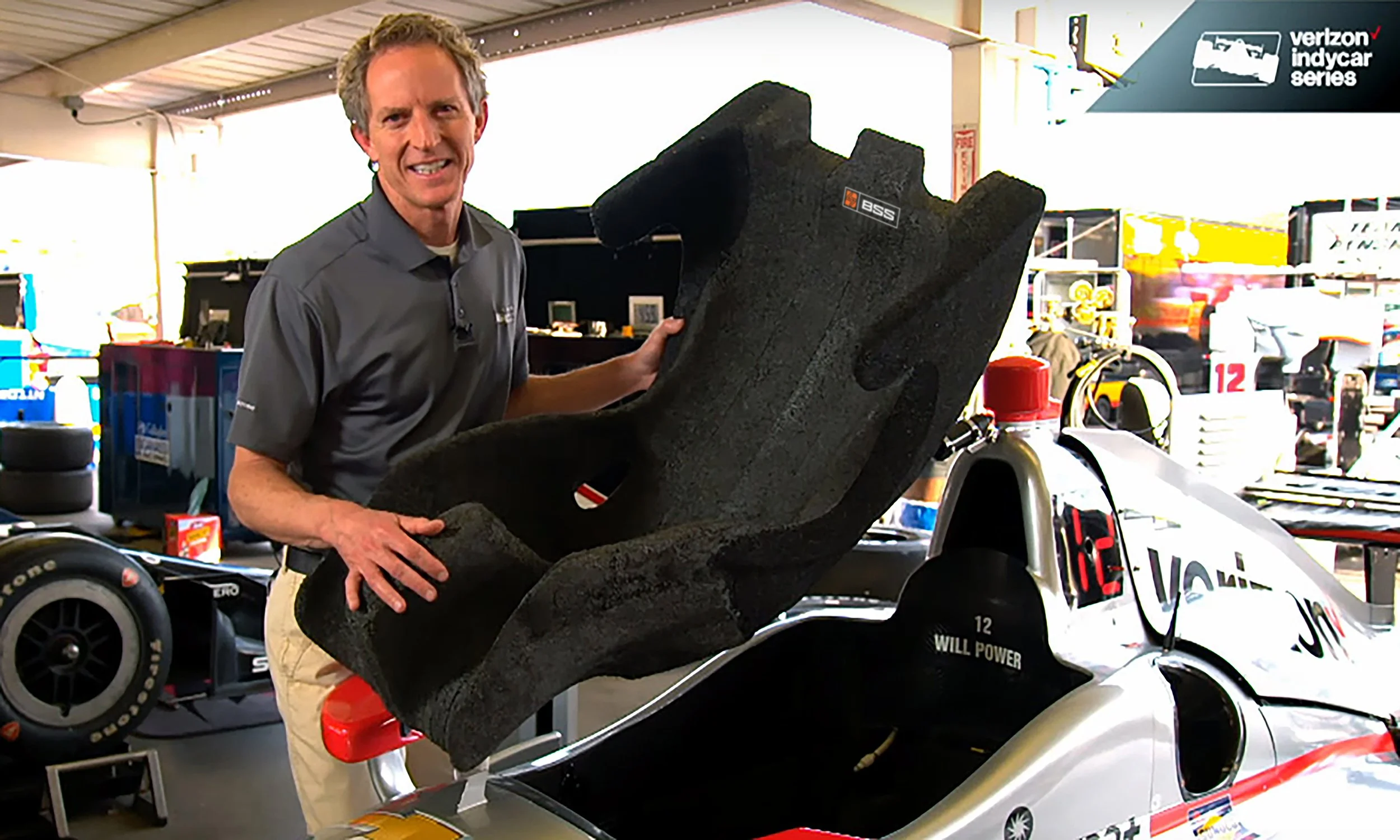 A man holding a Creapro foam racing seat in a garage or workshop, next to a race car with Verizon branding. The man is wearing a gray shirt and beige pants, and the background includes tires, tools, and various racing equipment.