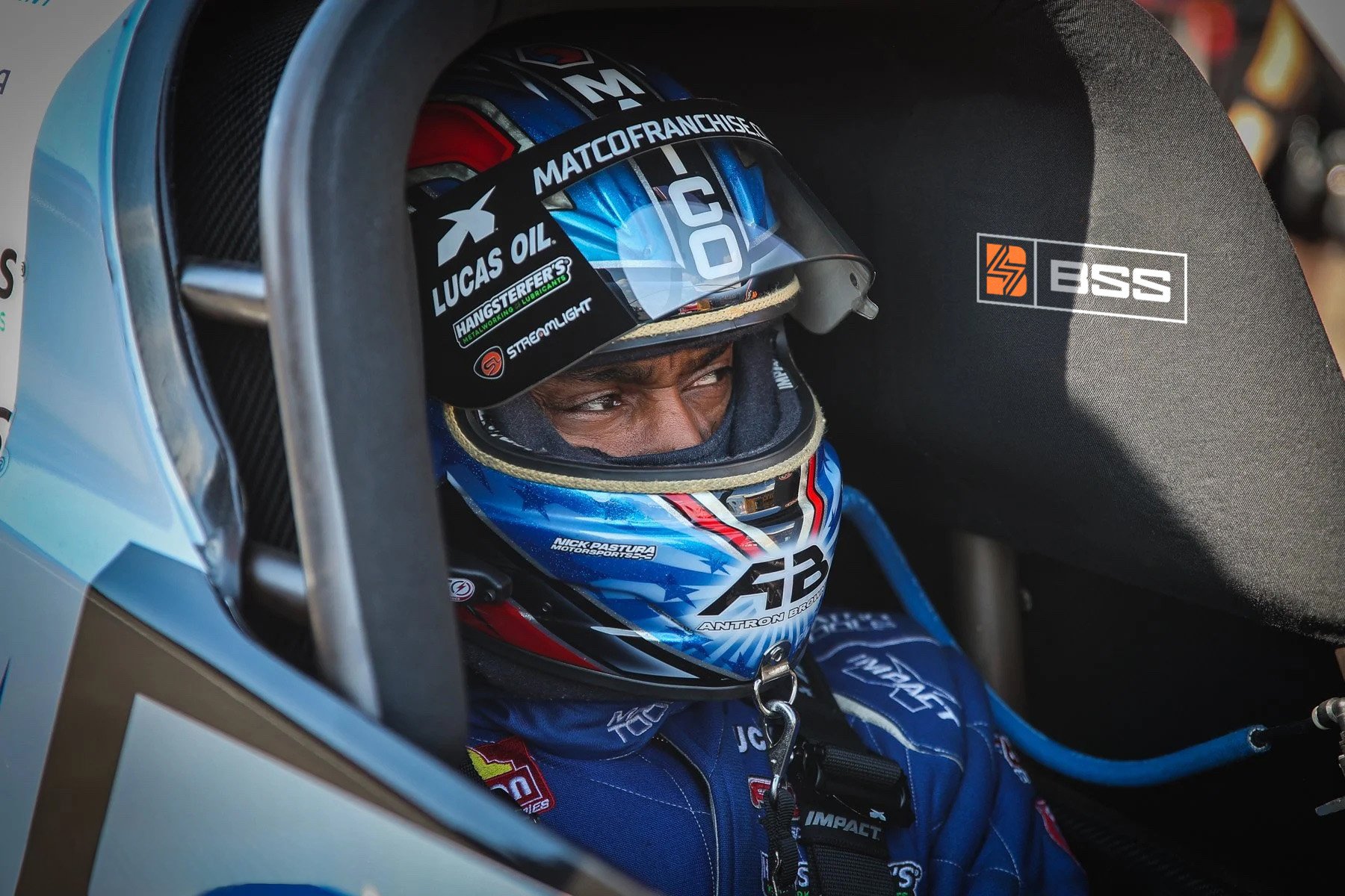 Close-up of a race car driver inside the cockpit, wearing a blue racing suit and helmet, with the helmet partially lifted showing the driver's eyes, surrounded by the car's interior.