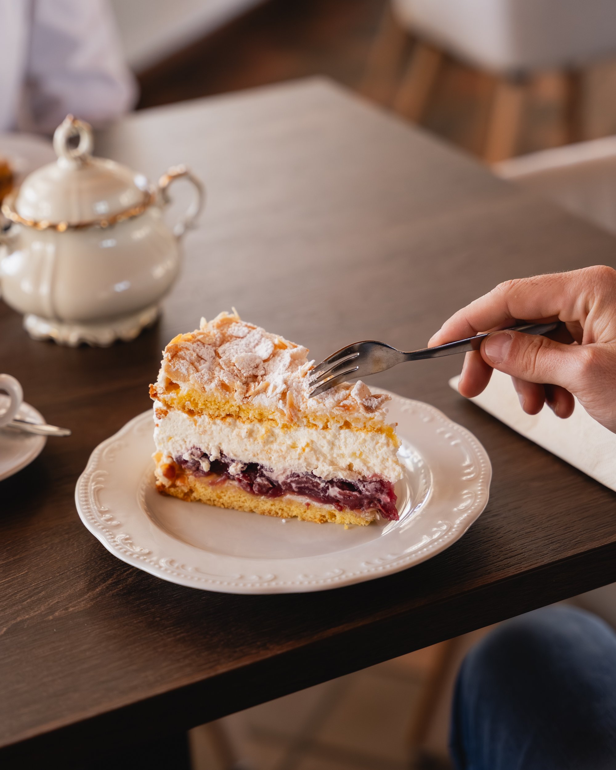 Ein Stück Käsekuchen mit Kirschen auf einem weißen Teller, neben einer Teekanne und einer Tasse auf einem Holztisch.