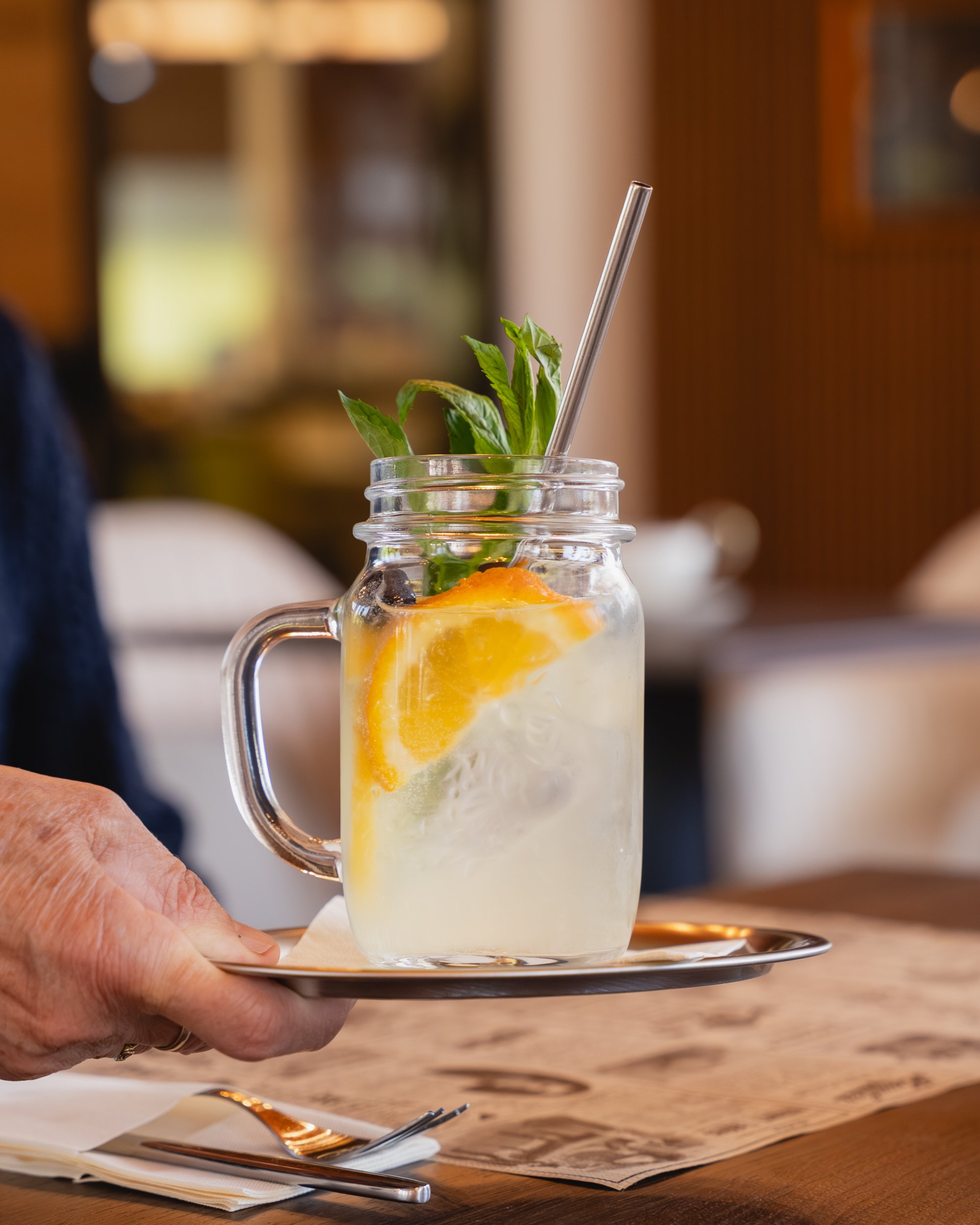 Eine Hand hält ein Glas Zitronenlimonande mit Zitronenscheiben, Minzblättern und Eiswürfeln, serviert auf einem kleinen silbernen Teller auf einem Holztisch.