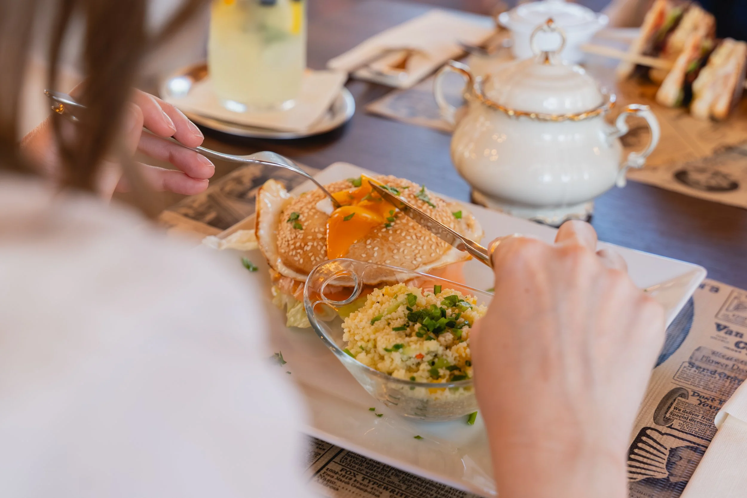 Person isst einen Hamburger mit Ei und Gemüse am Tisch, im Hintergrund Teekanne und Getränke, auf einem Tisch mit Papieren.