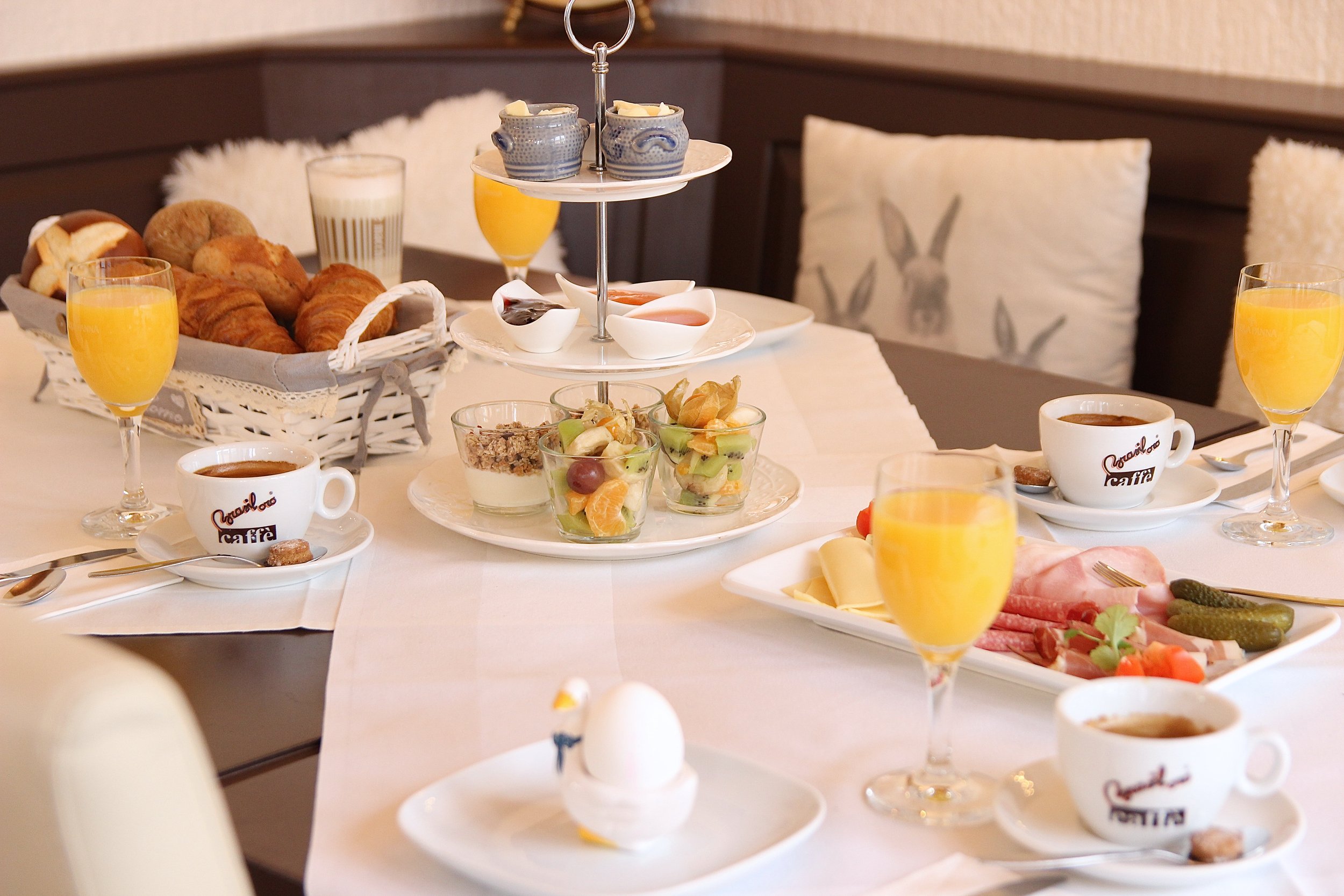 Frühstückstisch mit Säften, Croissants, Obst, Kaffee und Aufschnitt, serviert mit Tassen und Gläsern auf einer weißen Tischdecke.