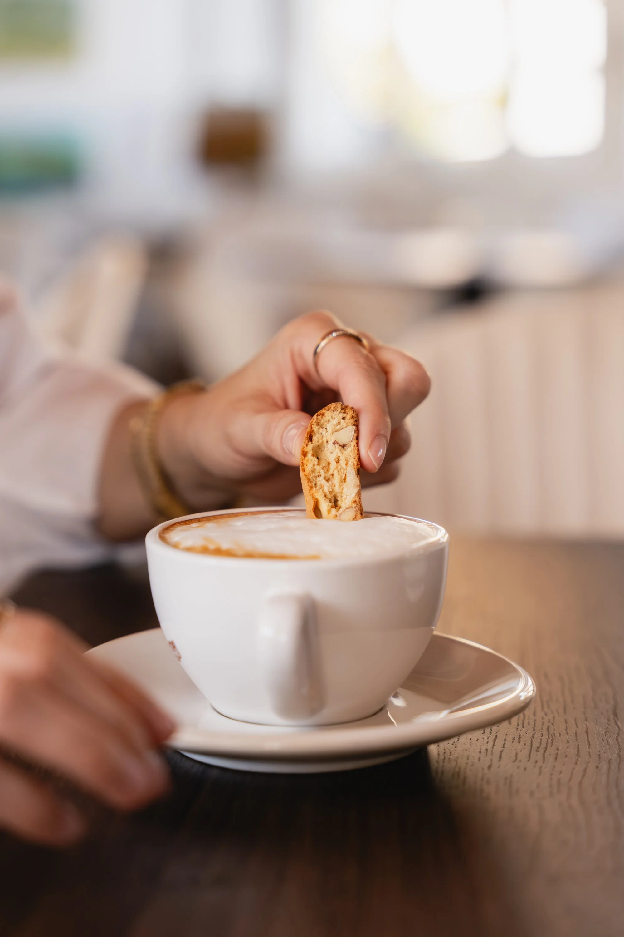 Eine Person taucht einen Keks in eine Tasse Kaffee oder Cappuccino. Die Hand der Person hält den Keks über die Tasse, die auf einem Unterteller steht. Die Umgebung ist unscharf,  aber zeigt eine gemütliche Café.