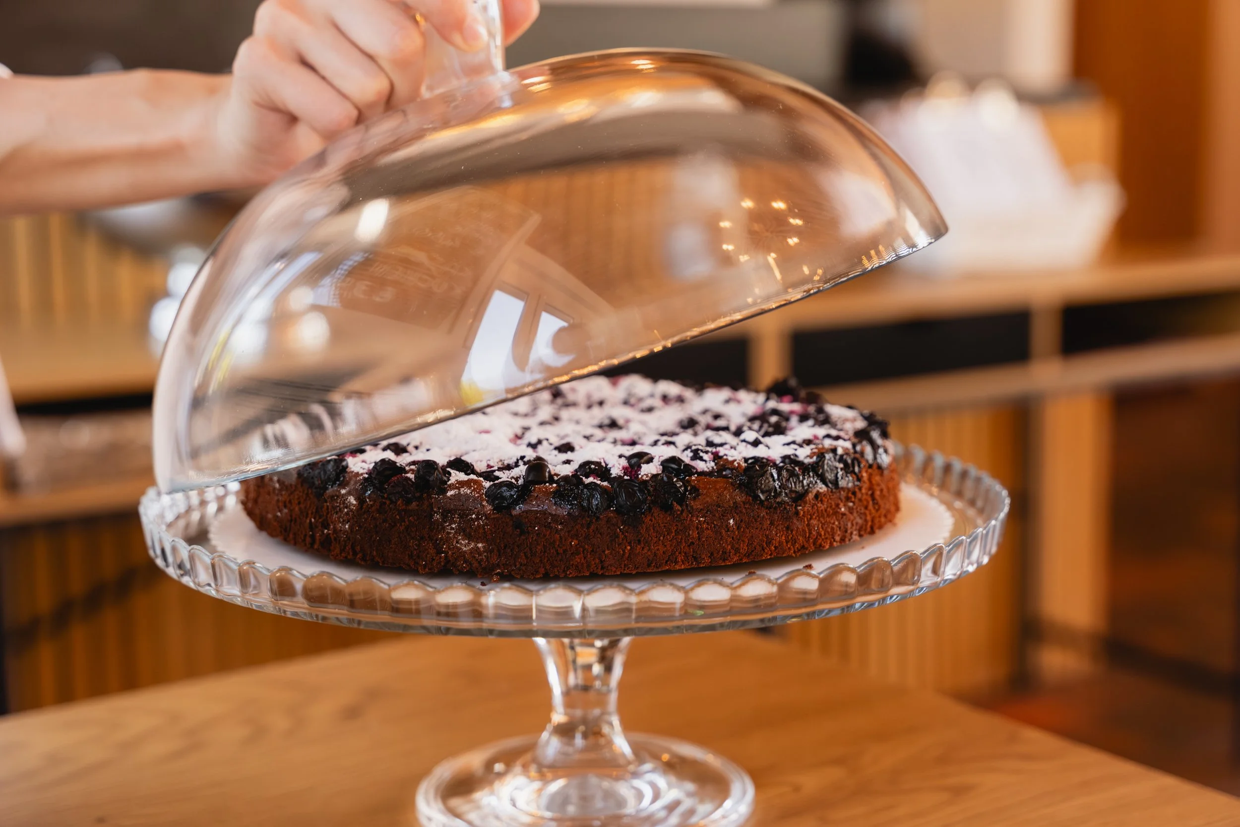 Ein Schokoladenkuchen mit Schokoladenstückchen und Puderzucker, der unter einer Glasabdeckung auf einem mehrstöckigen Tortenständer steht.