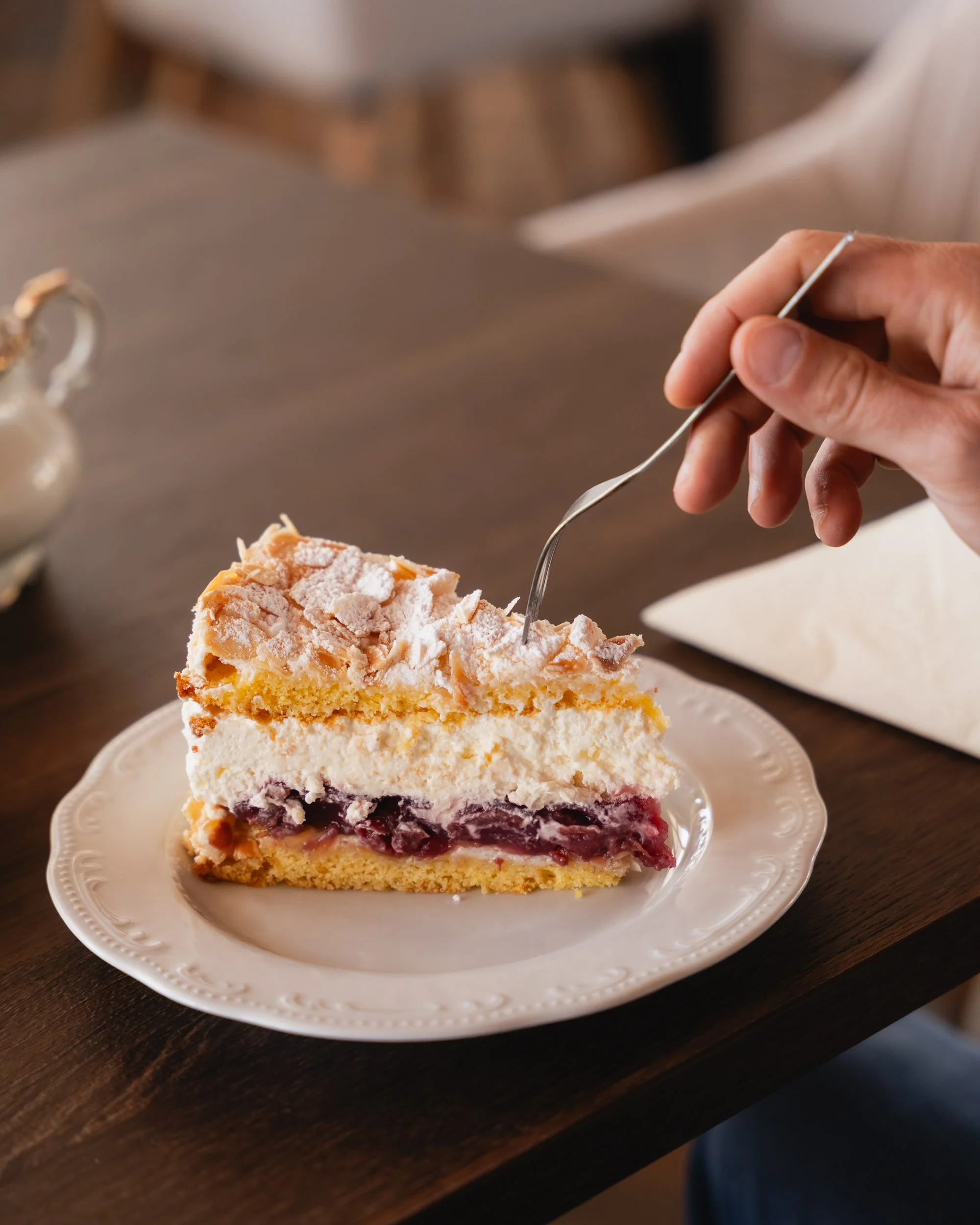 Ein Stück Kuchen mit Sahne, Pflaumen und Mandelblättchen auf einem weißen Teller auf einem Holztisch, im Hintergrund Tischdecke und Tasse.