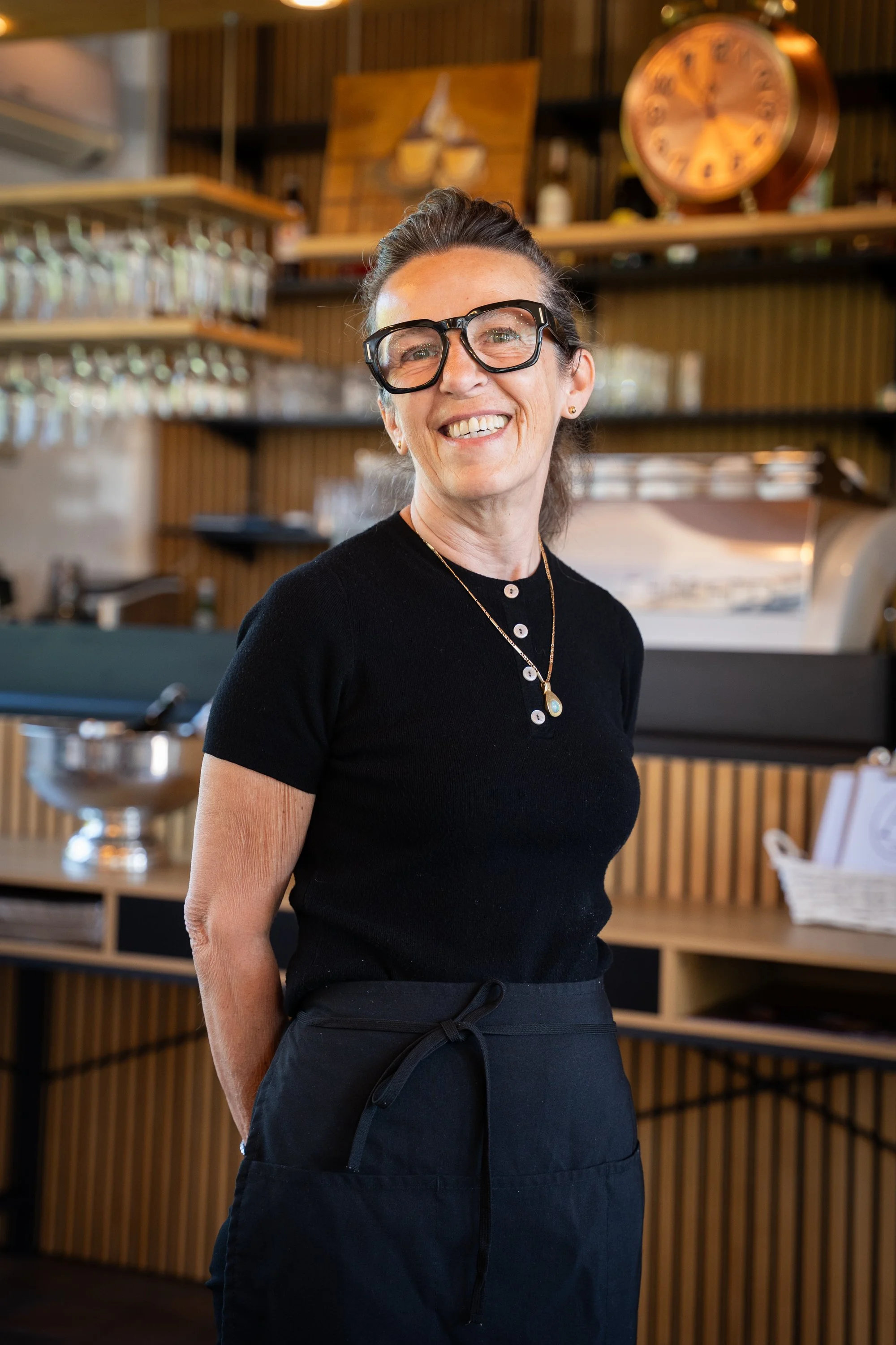 Eine lächelnde Frau mit Brille und dunklem Pullover, im Hintergrund eine Bar mit Weinregalen, einer Uhr und Dekorationen.