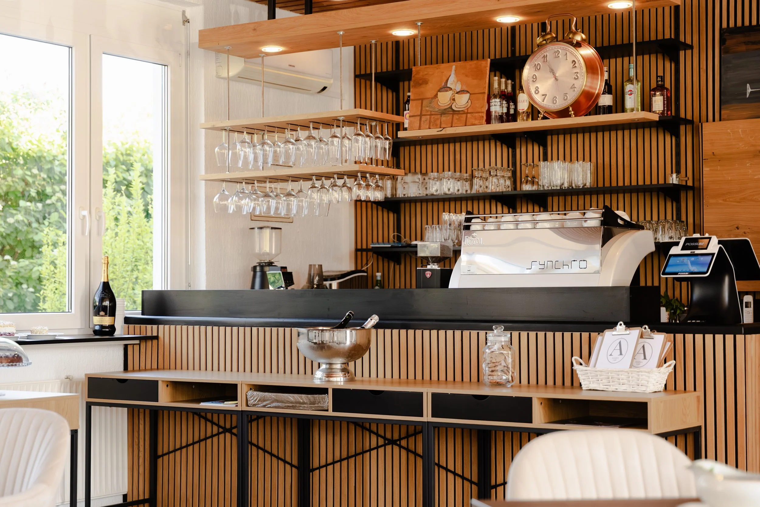 Innenraum eines modern eingerichteten Cafés oder Restaurants mit Holzregalen, einer Espresso-Maschine, Wein- und Spirituosenflaschen, hängenden Weingläsern, einem großen Wanduhren, einem Fenster mit Blick nach draußen und mehreren Tischen.