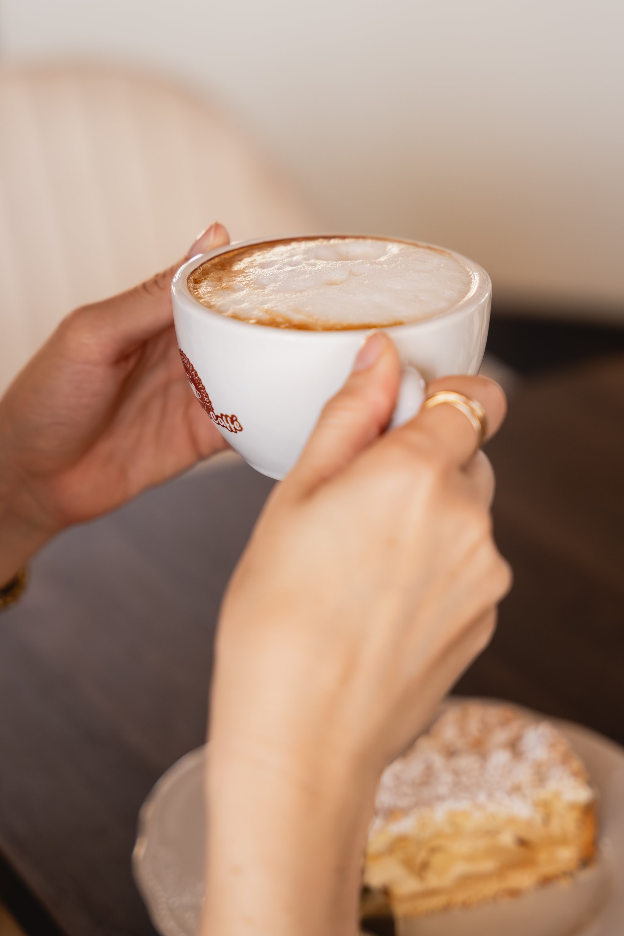 Person hält eine Tasse Cappuccino mit Milchschaum, im Hintergrund steht ein Teller mit Croissant.