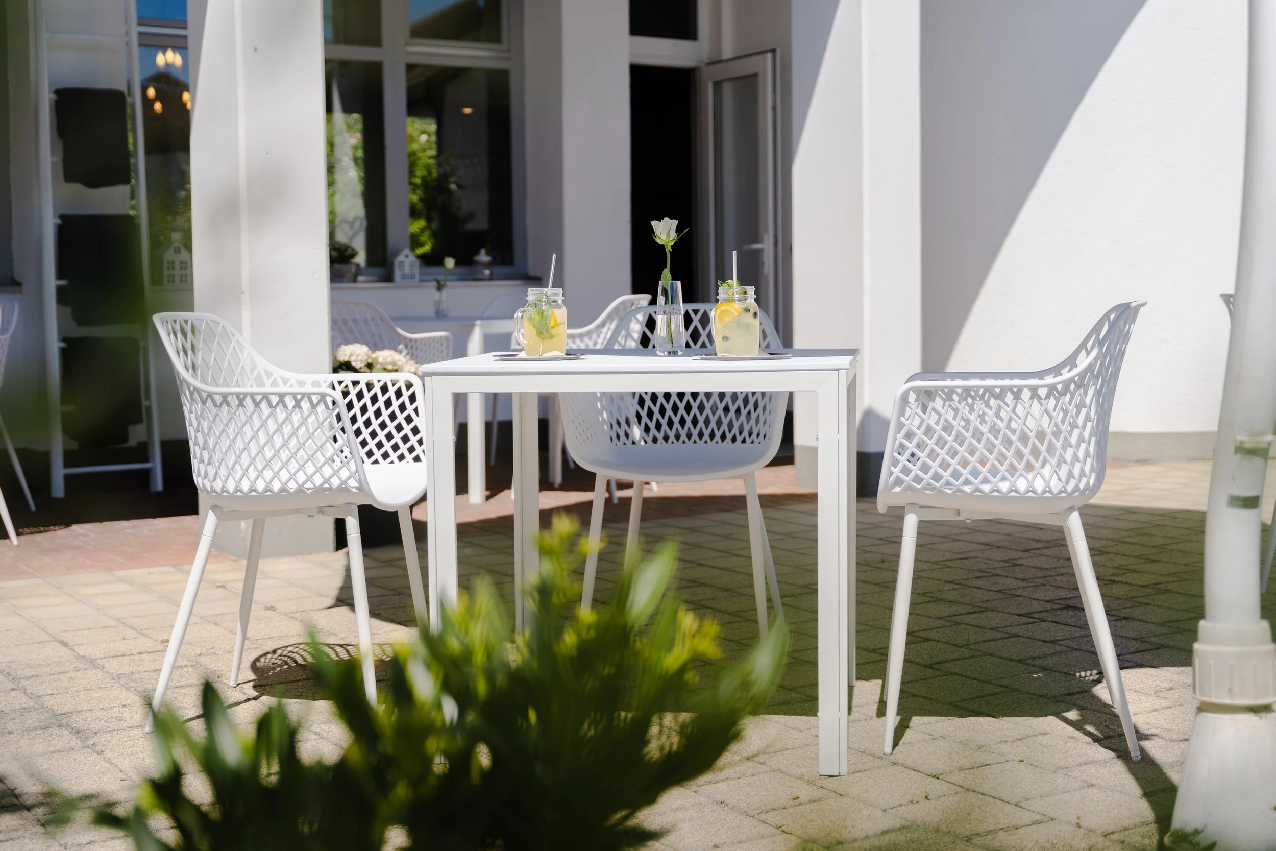 Ein weißer Tisch mit drei Gläsern Limonade, zwei mit Zitronen und eines mit einer weißblühenden Blume, auf einem Patio vor einem weißen modernen Haus.
