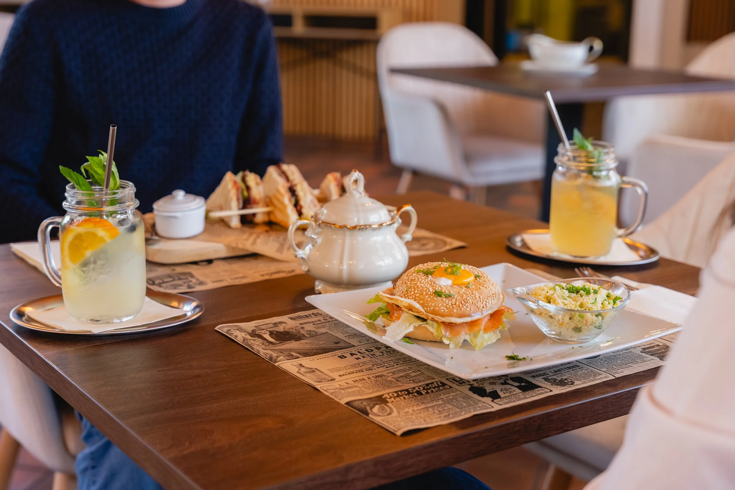 Ein Tisch im Café mit einem Sandwich, einem Burger, Salat, zwei Getränke in Gläsern mit Strohhalmen und Minzblättern. Im Hintergrund sind weiße Stühle und eine Holzverkleidung.