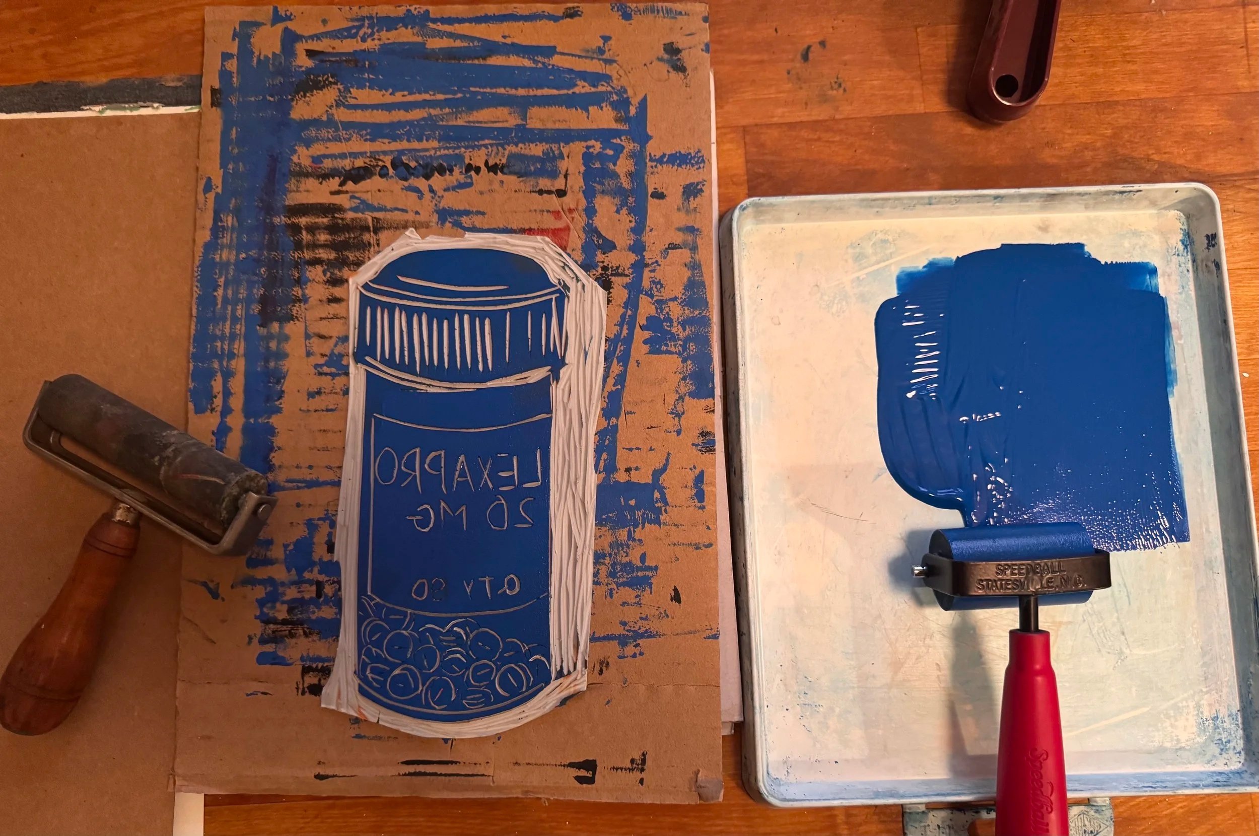A cardboard sign with a blue soda bottle illustration and text that reads '20 oz. LEXAPRO' is on a table. There is a tray with blue paint, a metal roller, a red handled roller, and a plastic cup of blue paint nearby. An LED flashlight is pointing at the sign, and part of a wooden mallet is visible on the table.