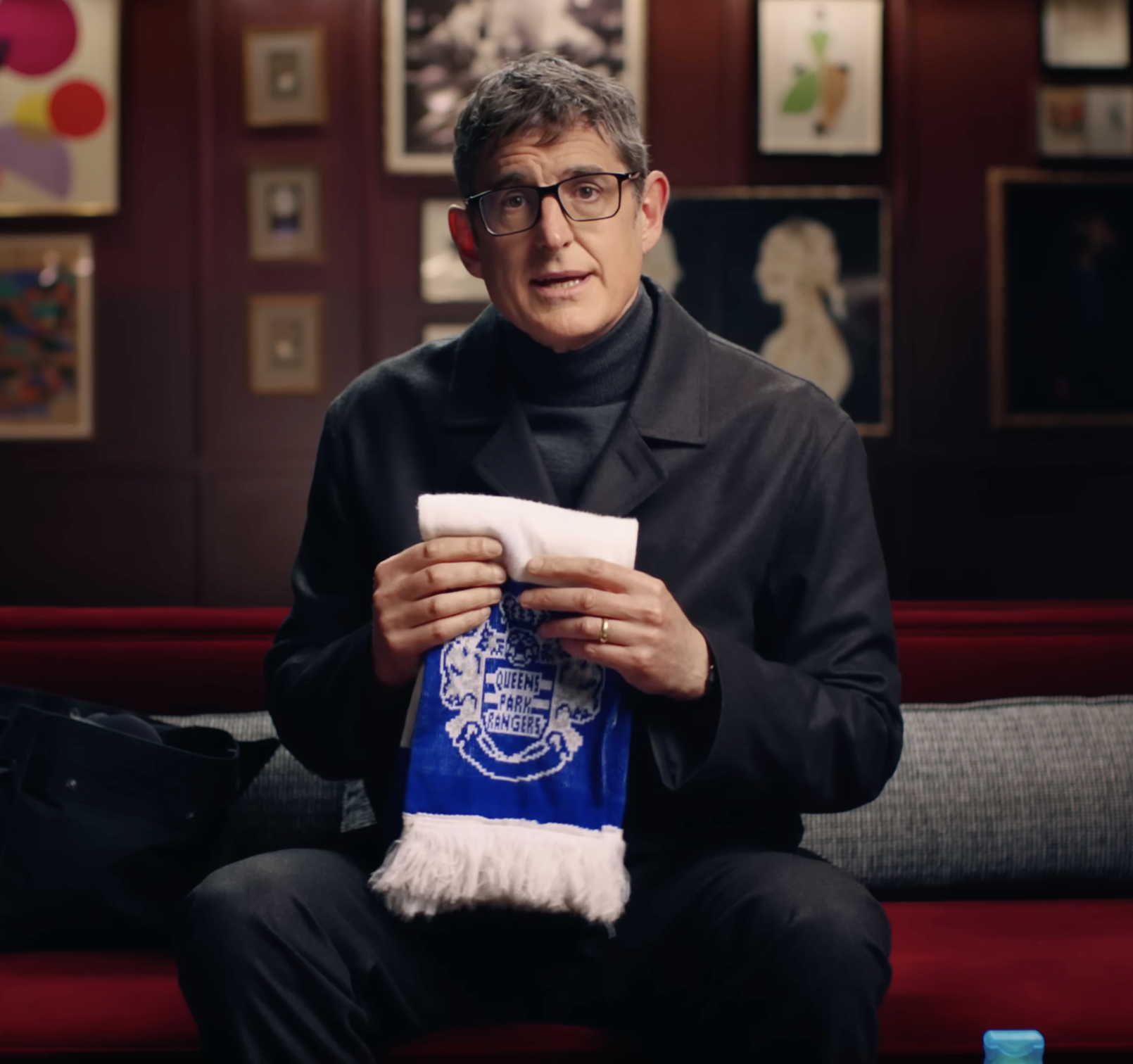 A man with glasses wearing a black coat sits on a red velvet bench, holding a Queens Park Rangers Christmas stocking, in front of a wall with framed artwork.