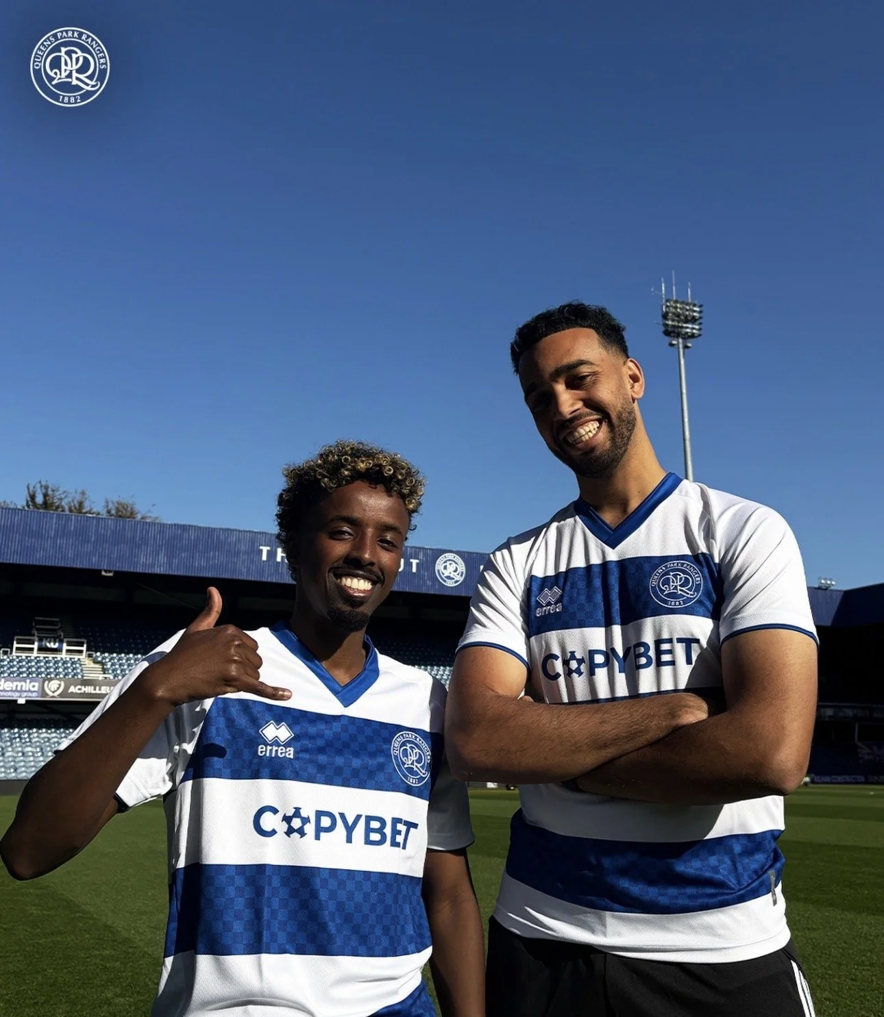 Two male soccer players in blue and white jerseys standing on a soccer field, smiling at the camera. One player on the left is making a phone gesture with his hand, the other on the right has arms crossed. Background shows stadium seating and a clear blue sky.
