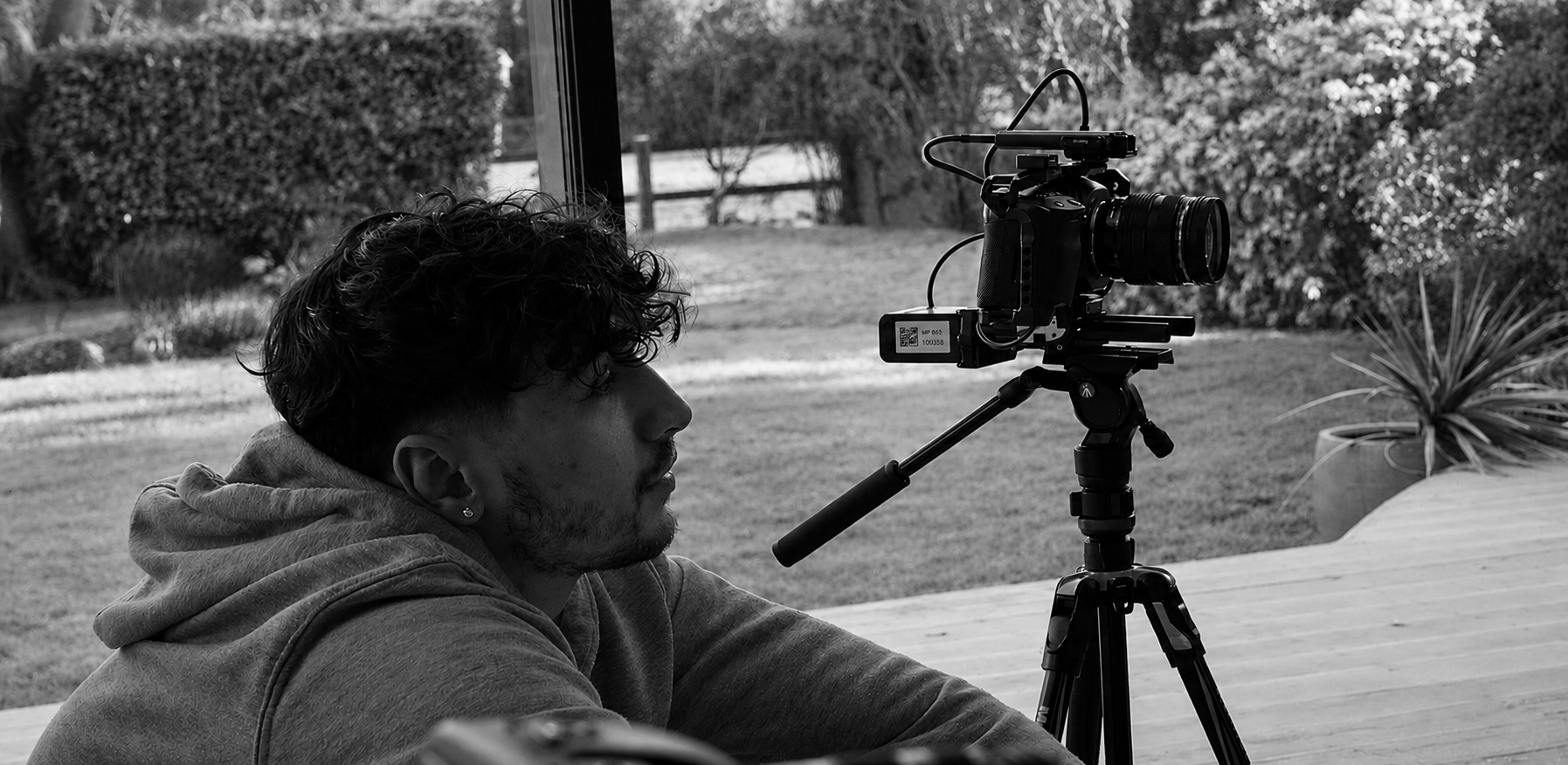 A person with curly hair wearing a hoodie sits at a table, looking at a camera mounted on a tripod, outdoors with a garden in the background.