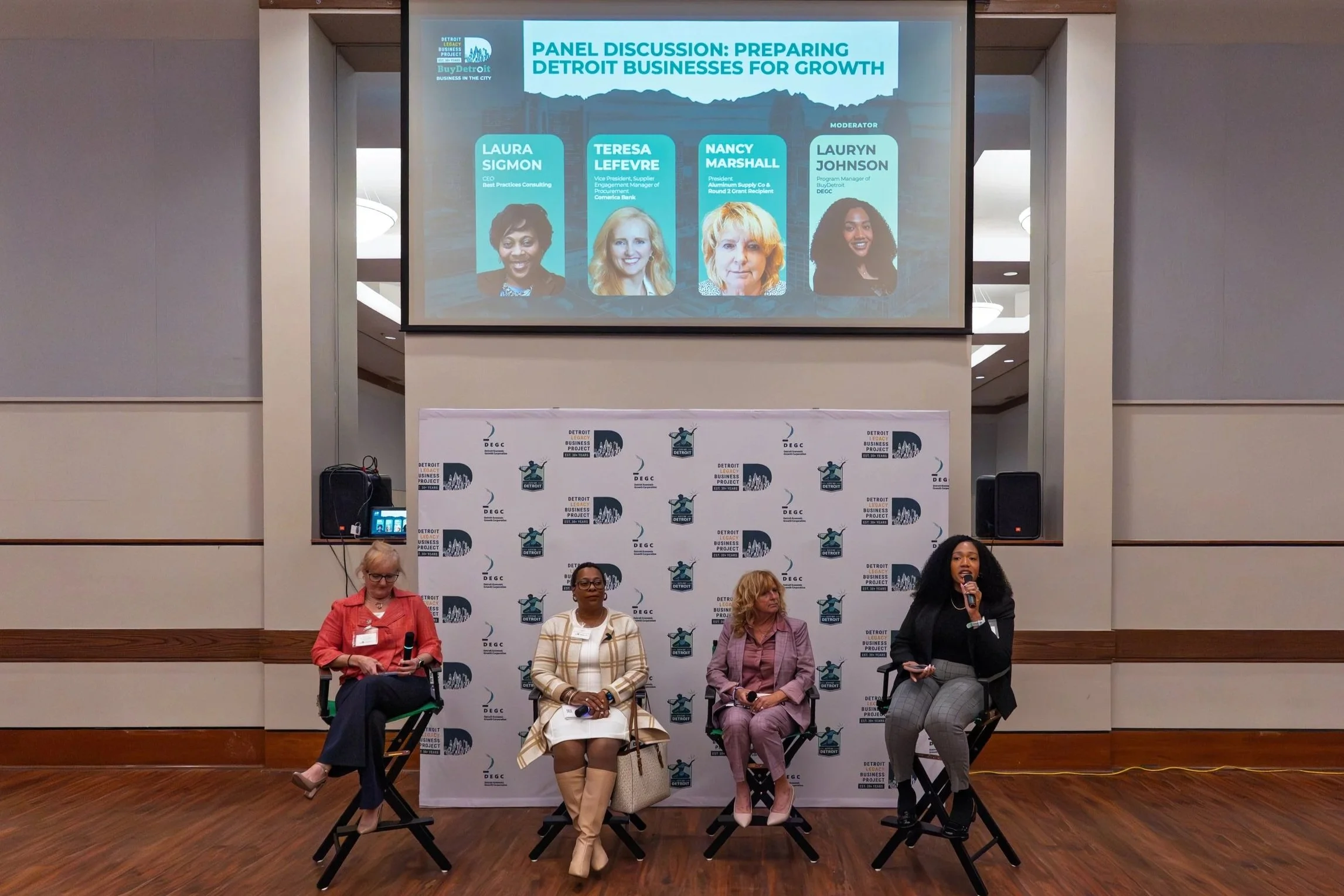 A panel discussion at a conference with four women seated on stage, each holding a microphone. A large screen behind them displays a title about preparing Detroit businesses for growth and features photos and names of the panelists, including Laura Sigmon, Teresa Lefevre, Nancy Marshall, and moderator Lauryn Johnson. The background includes a branded backdrop with logos of the Detroit Business Project.