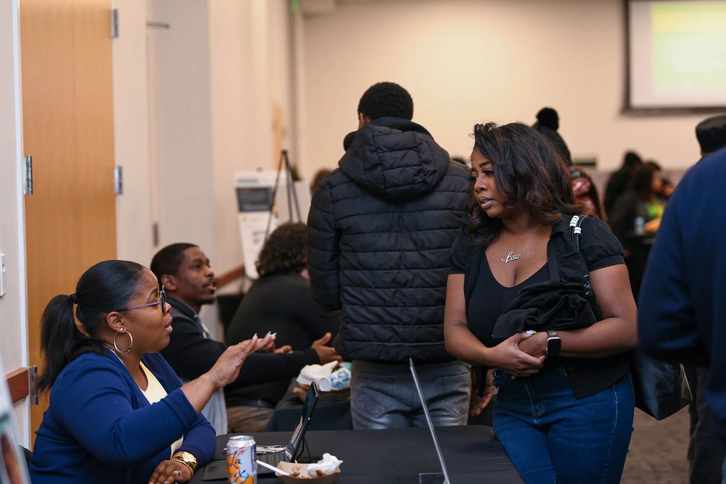 Two women sitting at a table and engaged in conversation during a professional event. One standing man with glasses and curly hair, wearing a navy jacket, is smiling and talking to the seated woman, who is smiling back and wearing glasses and a dark jacket. There are other women in the background, some working on laptops, with a blue banner that reads 'Grow Your Business Networking' behind them.
