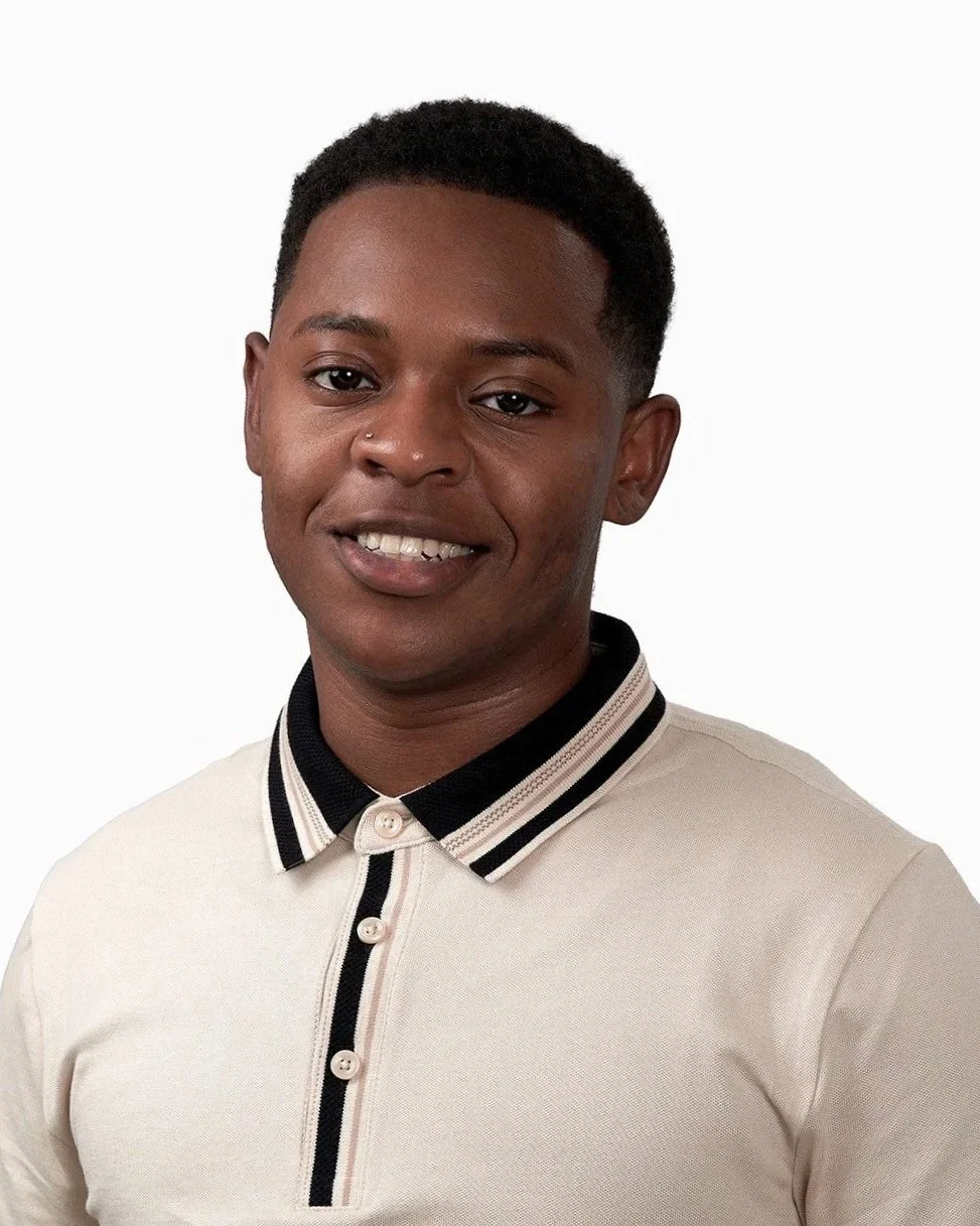 A young man with short curly hair, smiling, wearing a beige polo shirt with black and white striped collar and placket, standing against a plain white background.