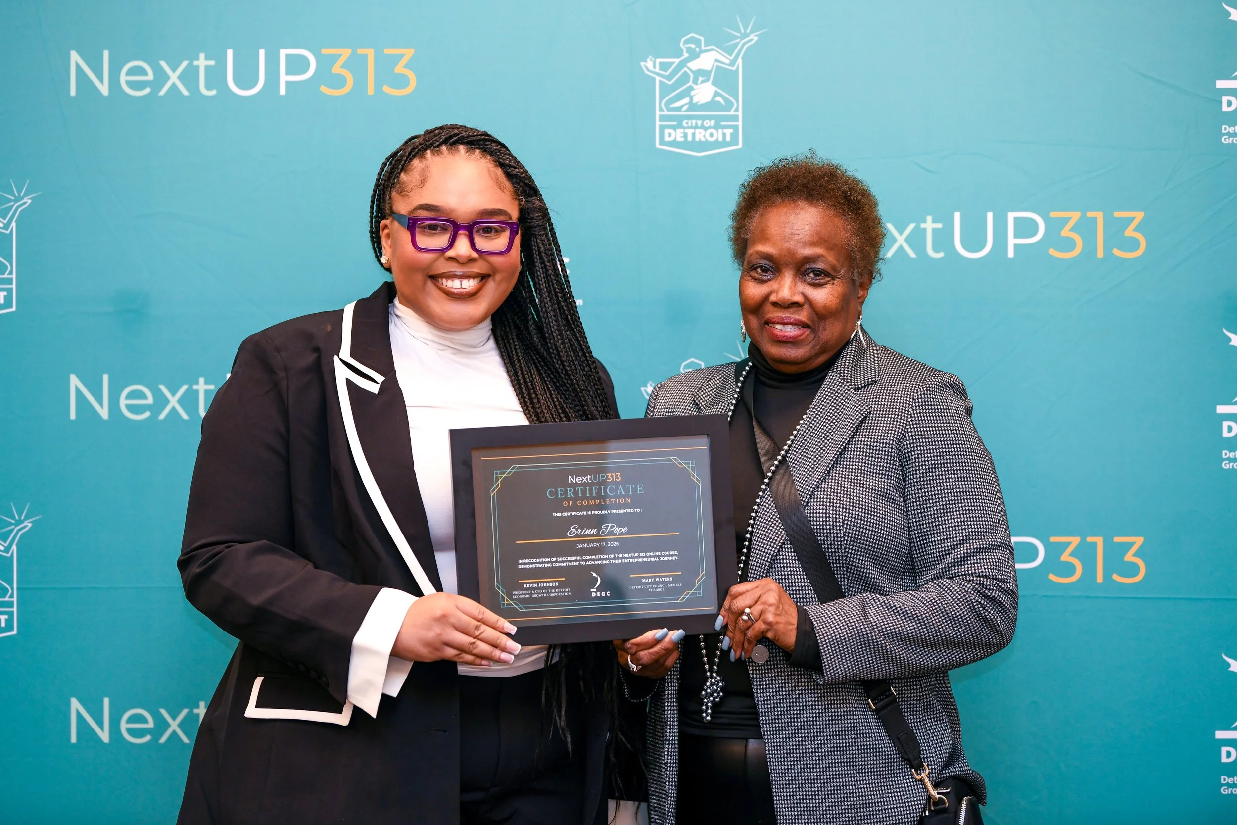 Two women standing in front of a blue backdrop with text and logos, holding a framed certificate of completion, smiling for the camera.