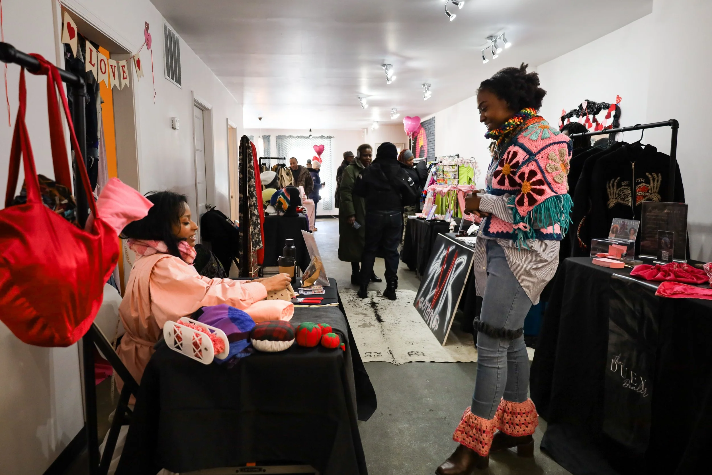 People shopping at a craft fair or boutique, browsing clothes and handmade items, with heart-shaped balloons and Valentine decorations in the background.