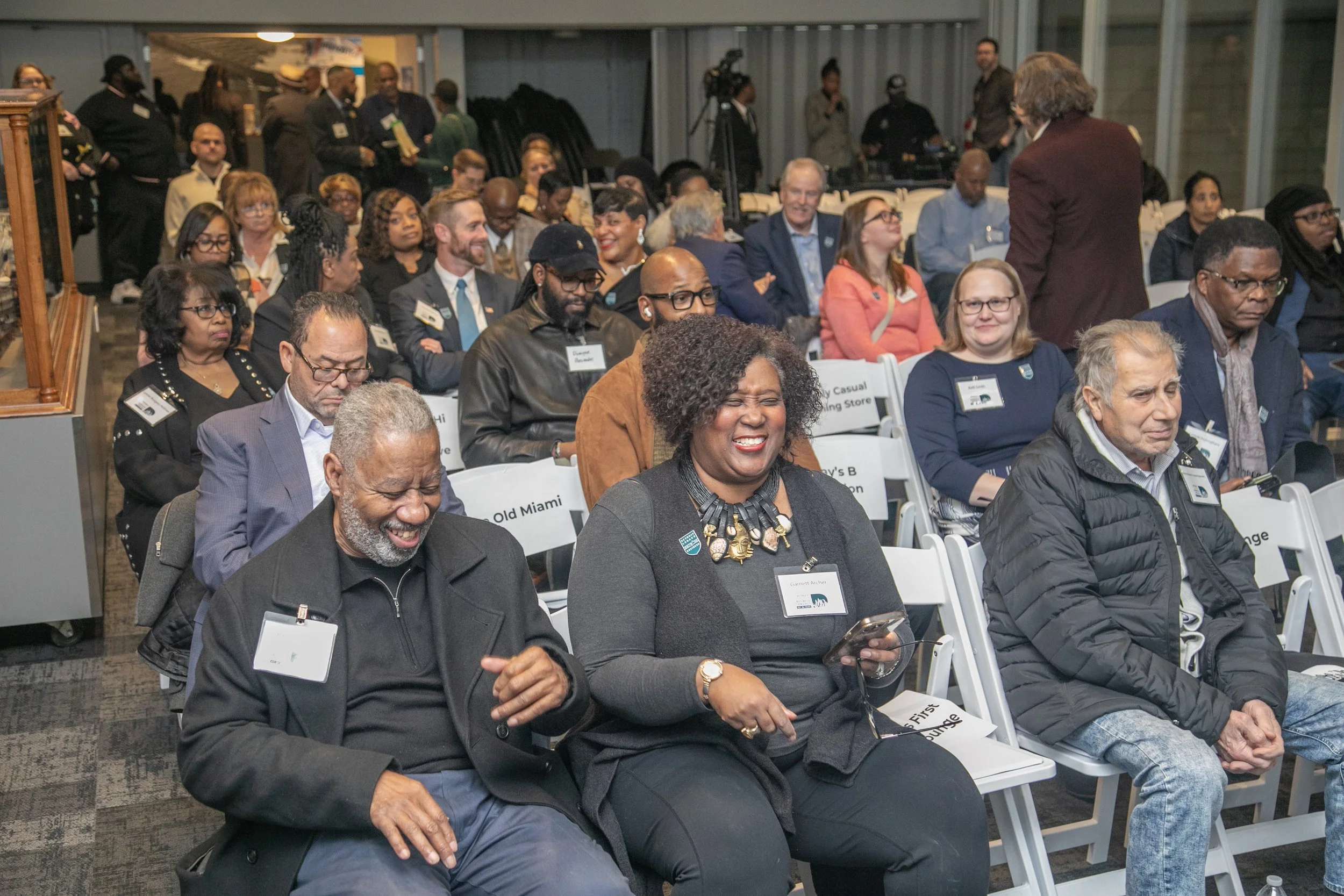 A diverse group of people seated in an indoor conference or event setting, some are smiling and engaging, with a woman in the front wearing a necklace and reading glasses. There are people with name tags, and some are looking at their phones or listening attentively.