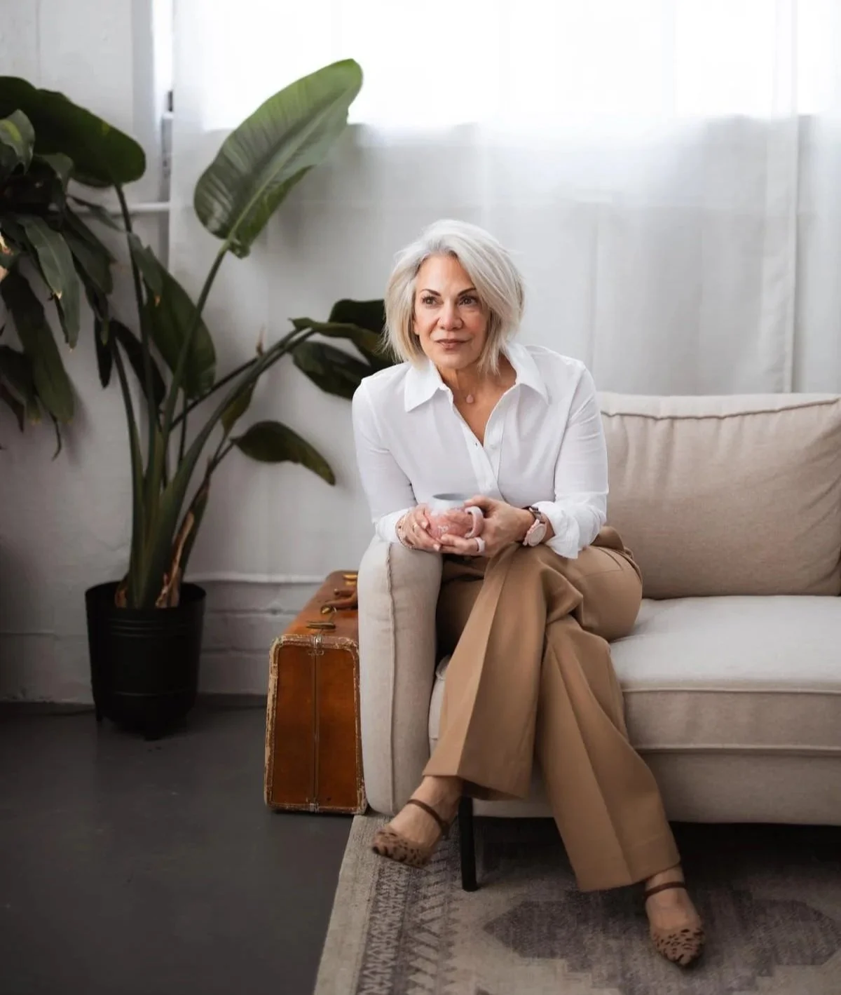 An older woman with gray hair, wearing a white blouse and tan pants, sits on a beige sofa holding a mug. There is a large green plant in a black pot to her left, a vintage suitcase beneath her, and a light-colored rug on the floor. The background includes white curtains.