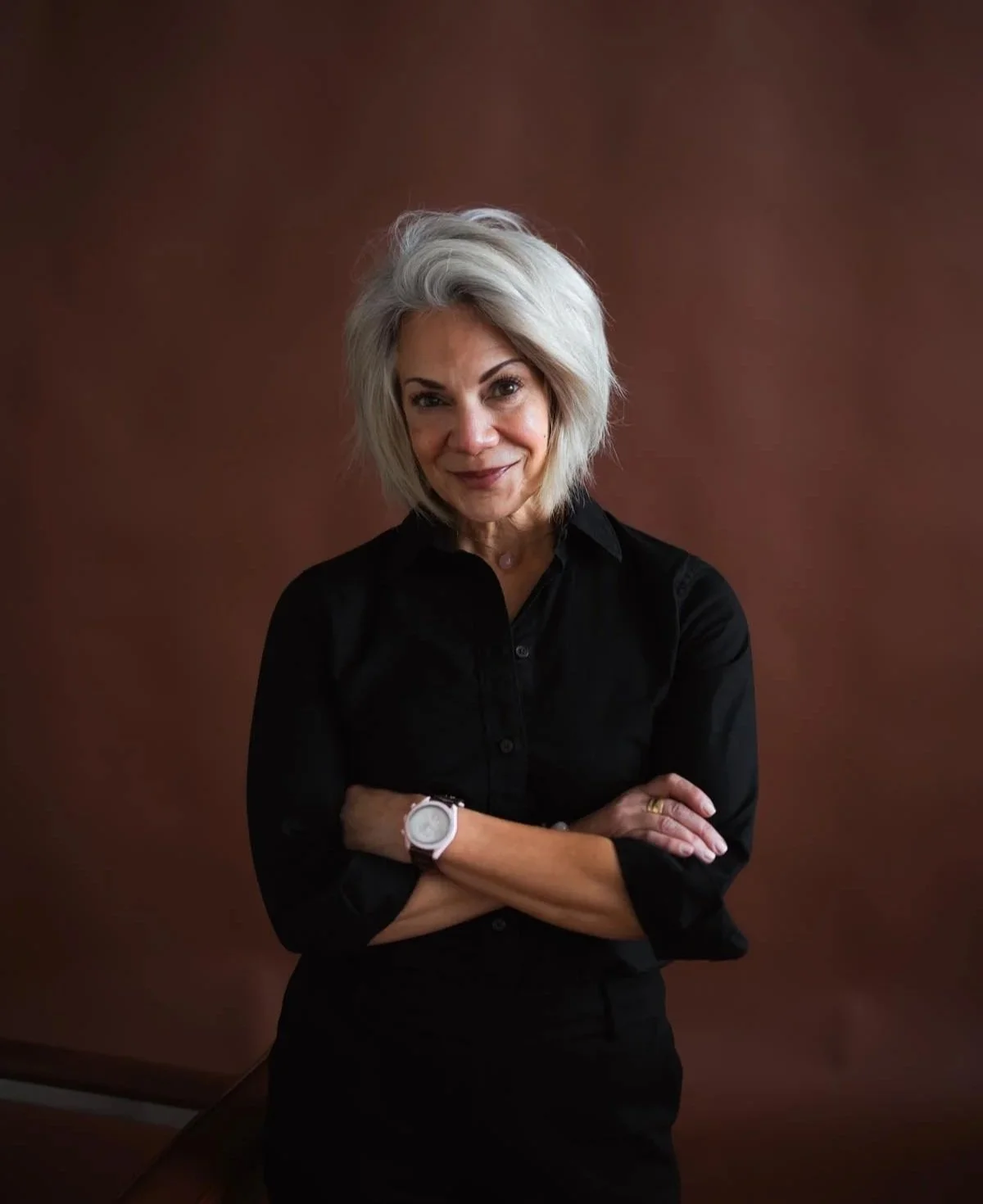 A woman with short, gray hair wearing a black shirt, standing with her arms crossed in front of a brown background, smiling at the camera.
