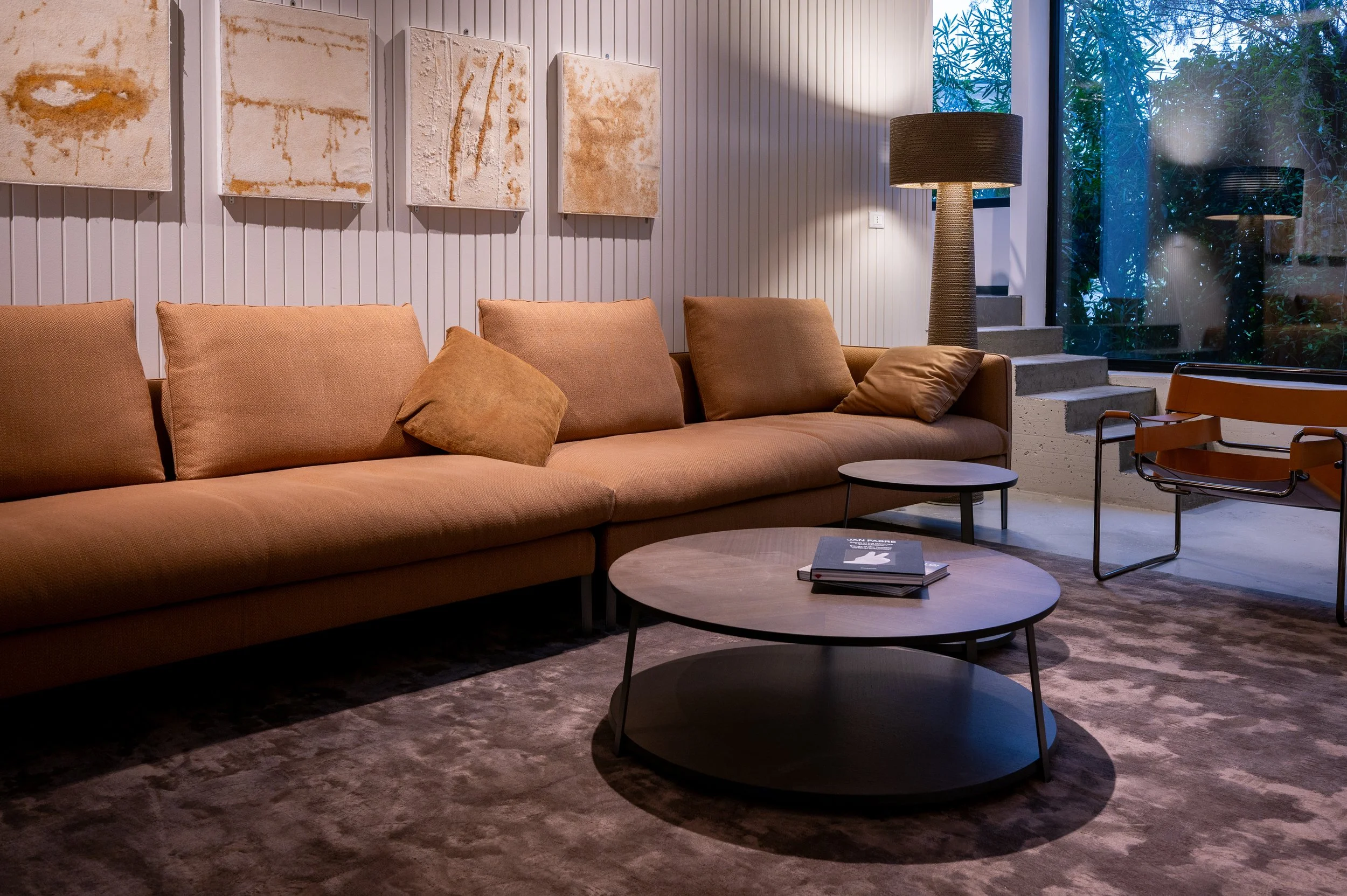 Interno di una sala di soggiorno con divano color terra, mensole con quadri, una grande lampada a stelo, un tavolino rotondo con libri e una grande finestra con vista su piante verdi.